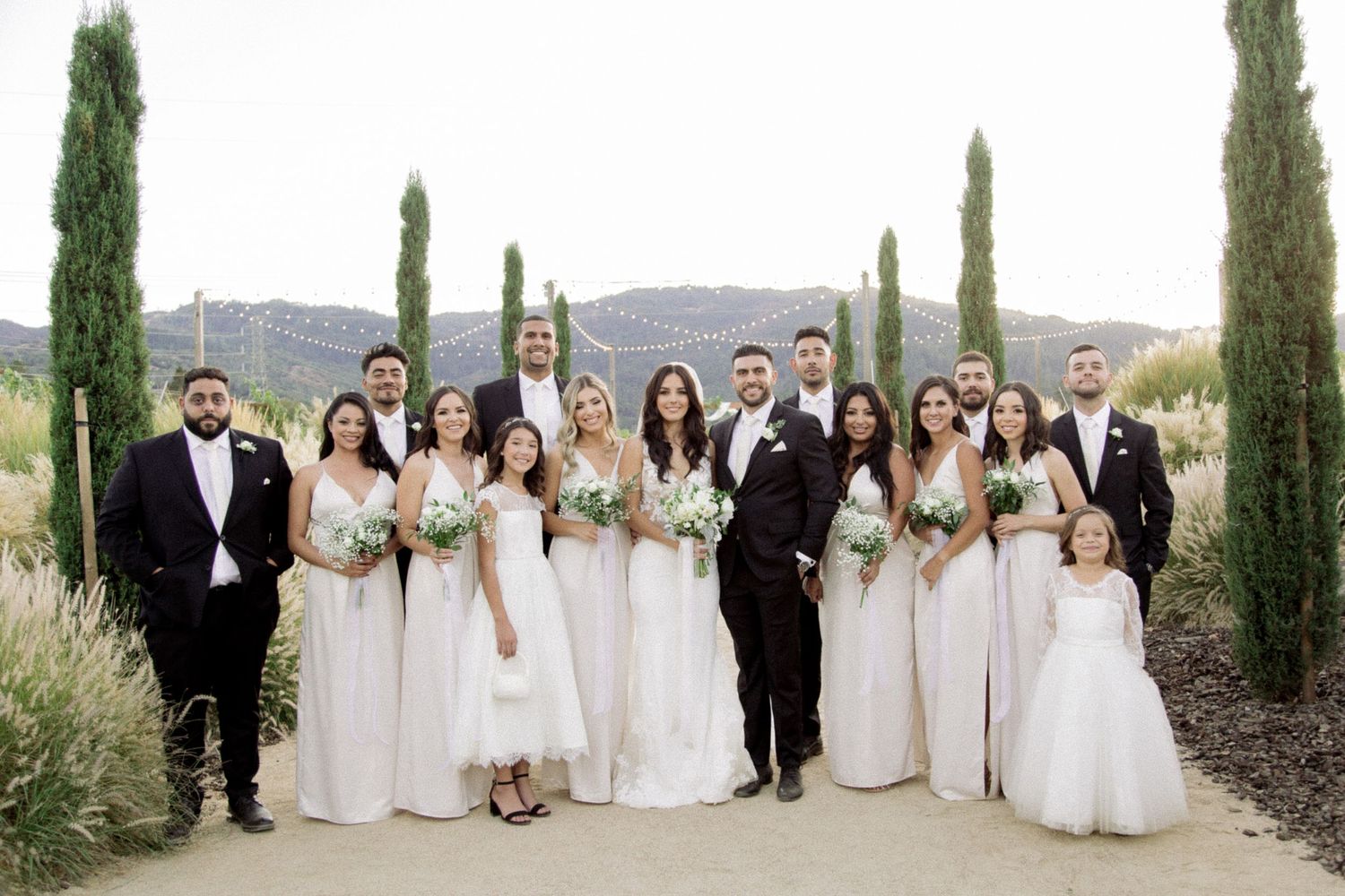 Wedding party in white dresses and black suits posed along cypress-lined path.