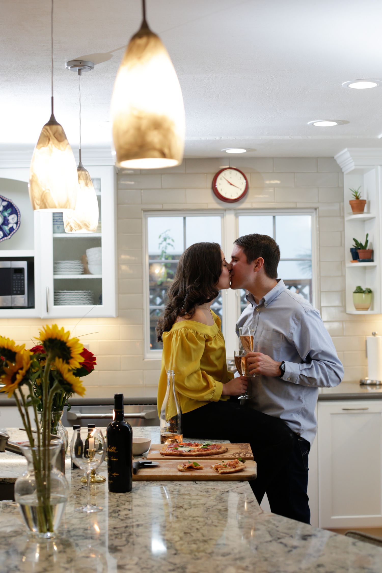 Romantic couple sharing wine and pizza in a bright modern kitchen with sunflowers on the counter.