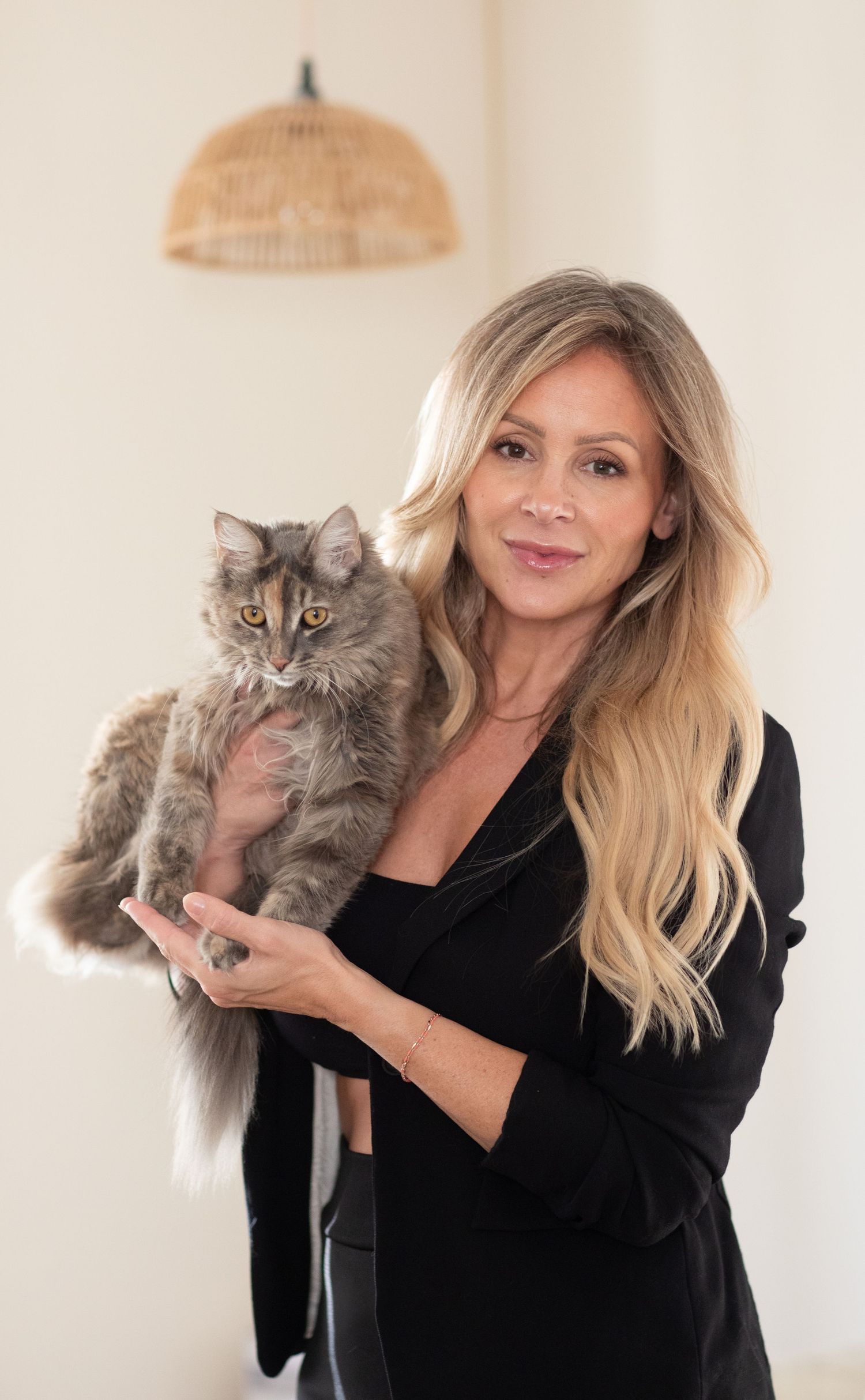 A woman in a black blazer holds a fluffy grey cat with blue eyes while posing in front of a woven pendant light.