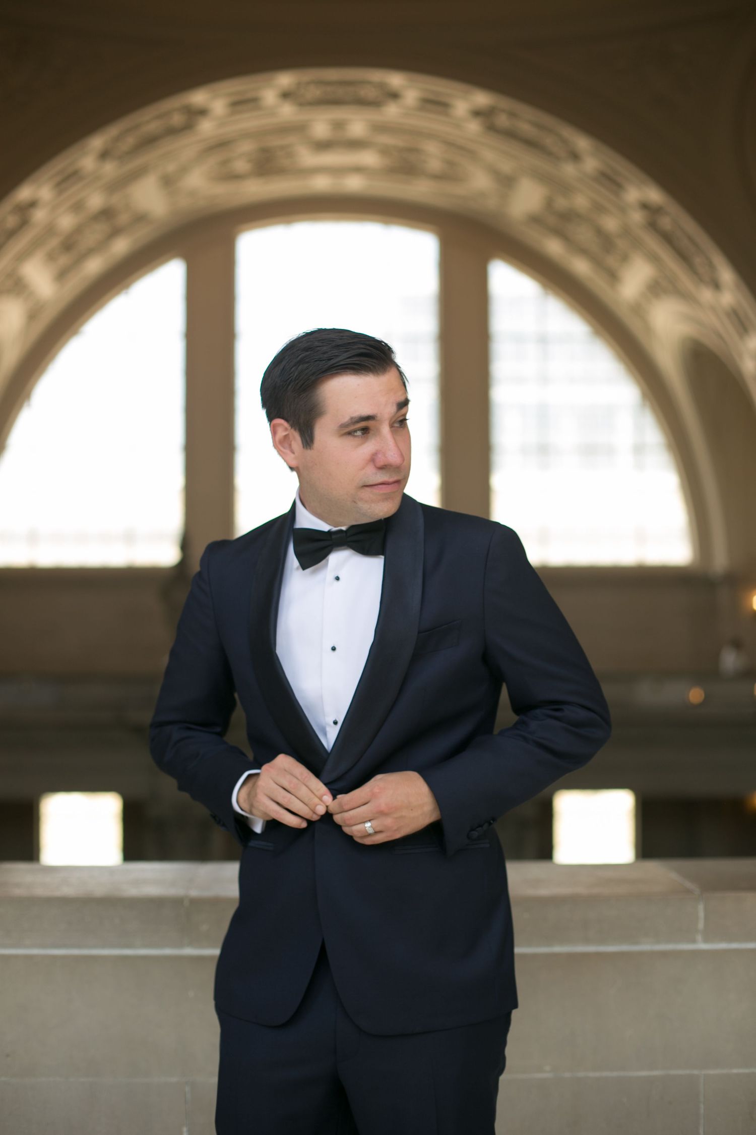 Person in classic black tuxedo standing beneath ornate archway.