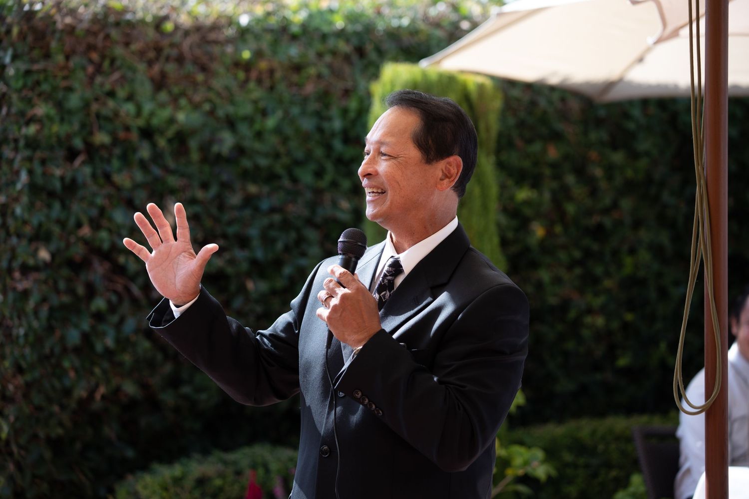 A person in a suit gestures while speaking at an outdoor wedding reception.