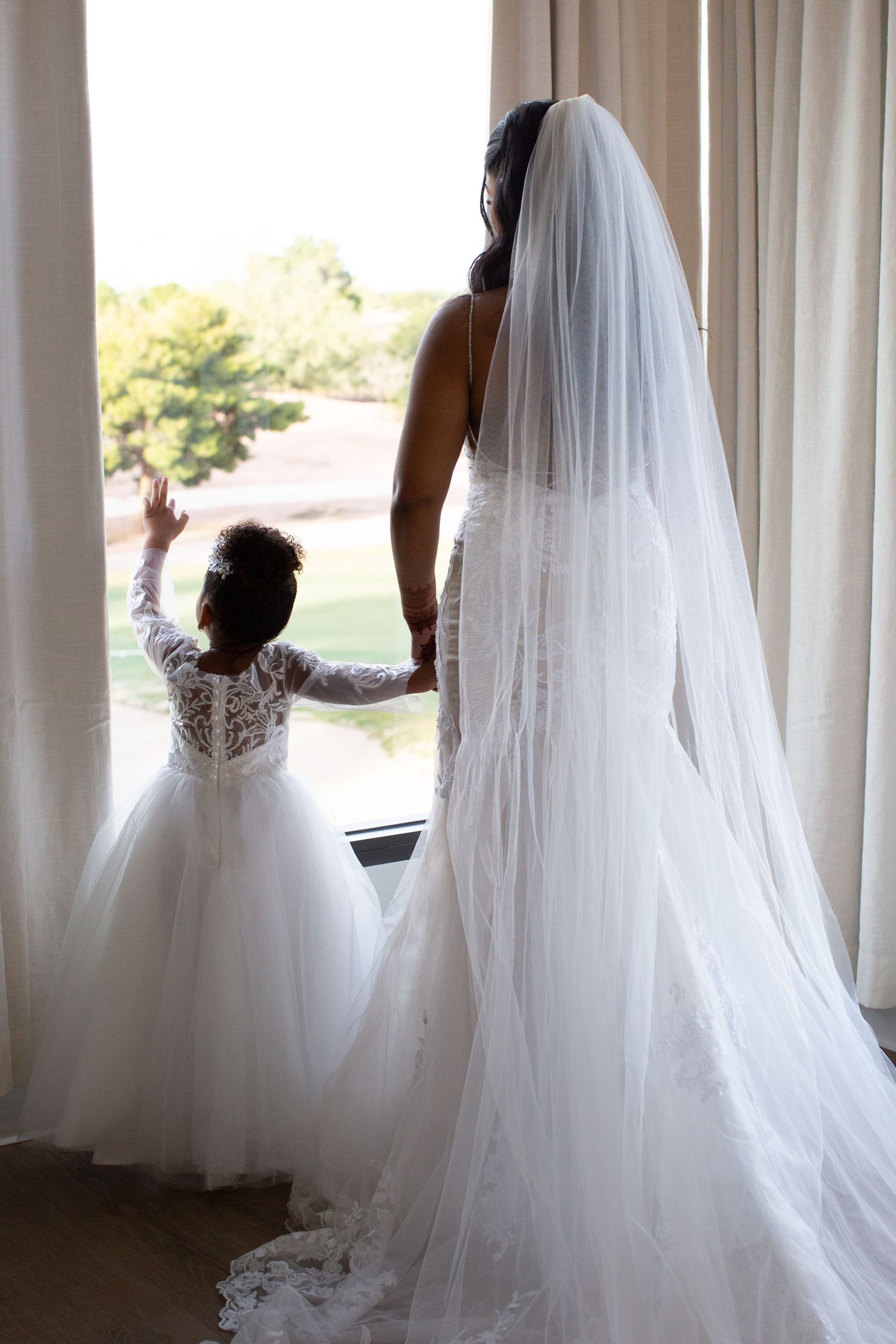 A bride in a flowing white gown and veil stands at a window with a small flower girl in a lace dress.