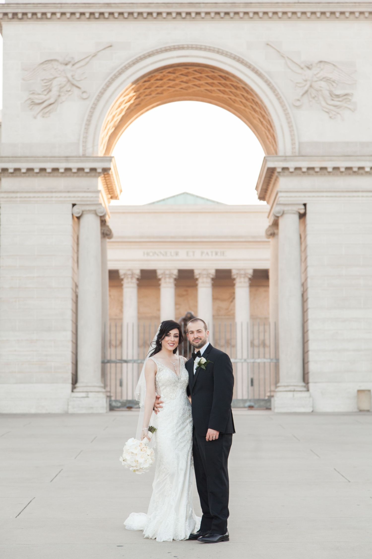 Couple poses for wedding photo beneath majestic stone archway with ornate architectural details.