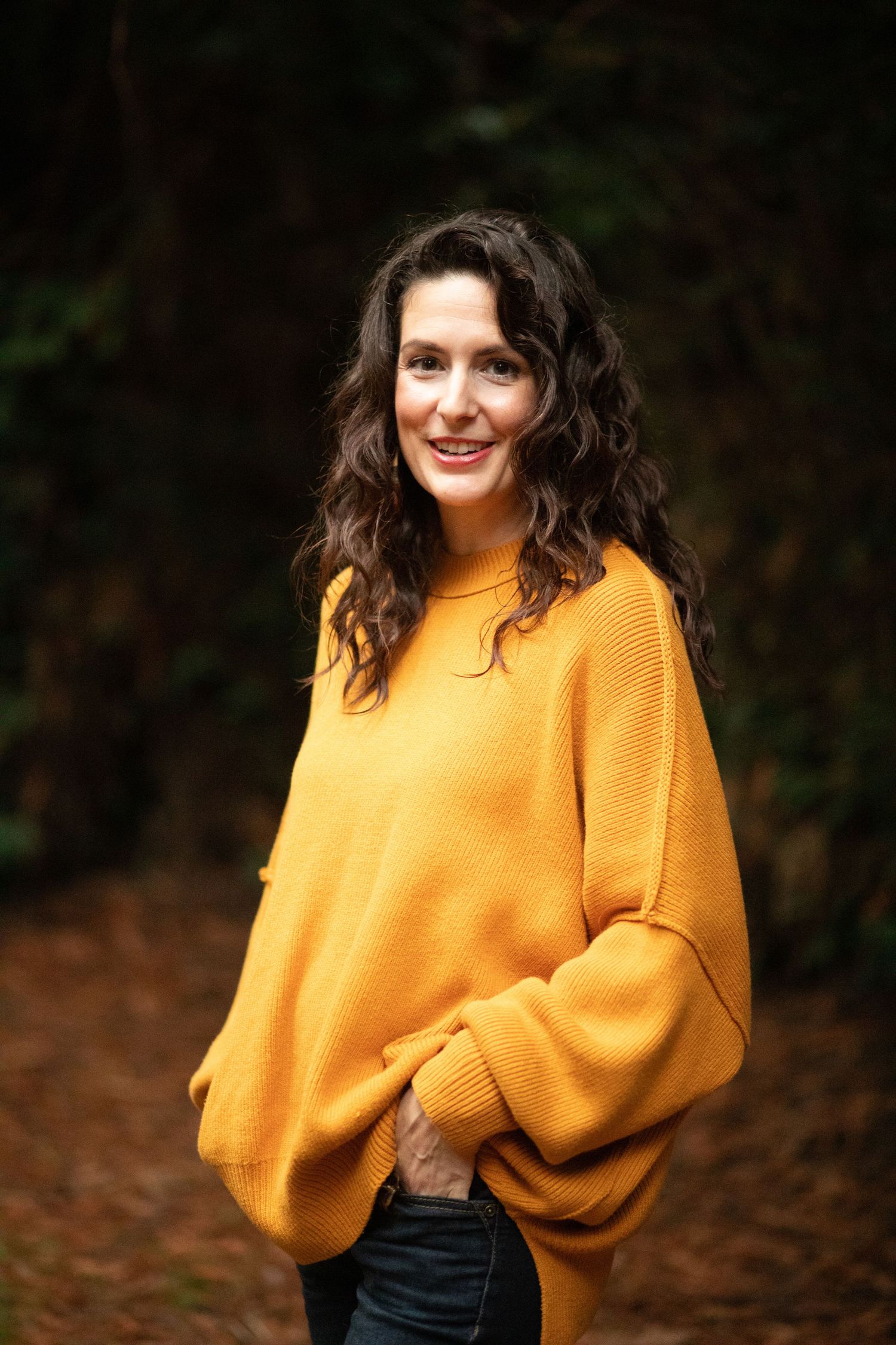 Portrait of person in oversized yellow sweater against dark forest background.