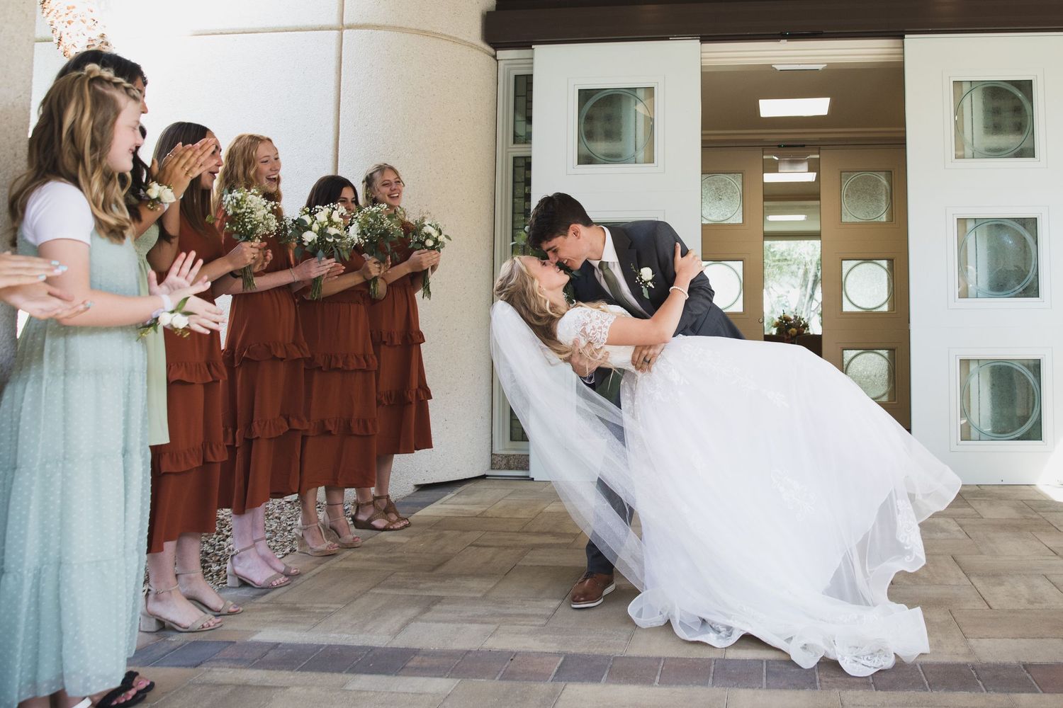 Wedding party watching romantic dip kiss between newlyweds at church entrance.