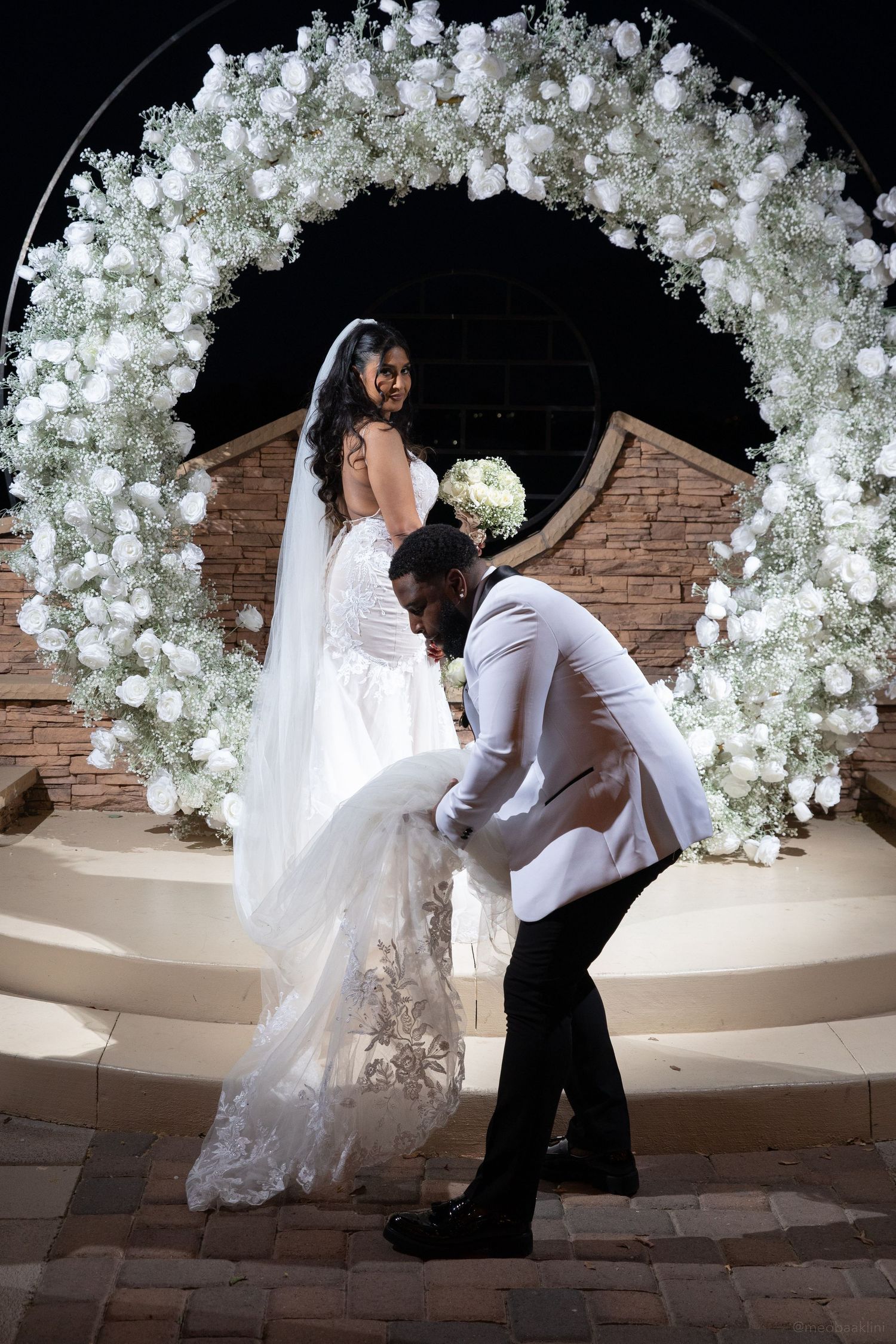 Romantic moment under a white floral arch as dress train is adjusted.
