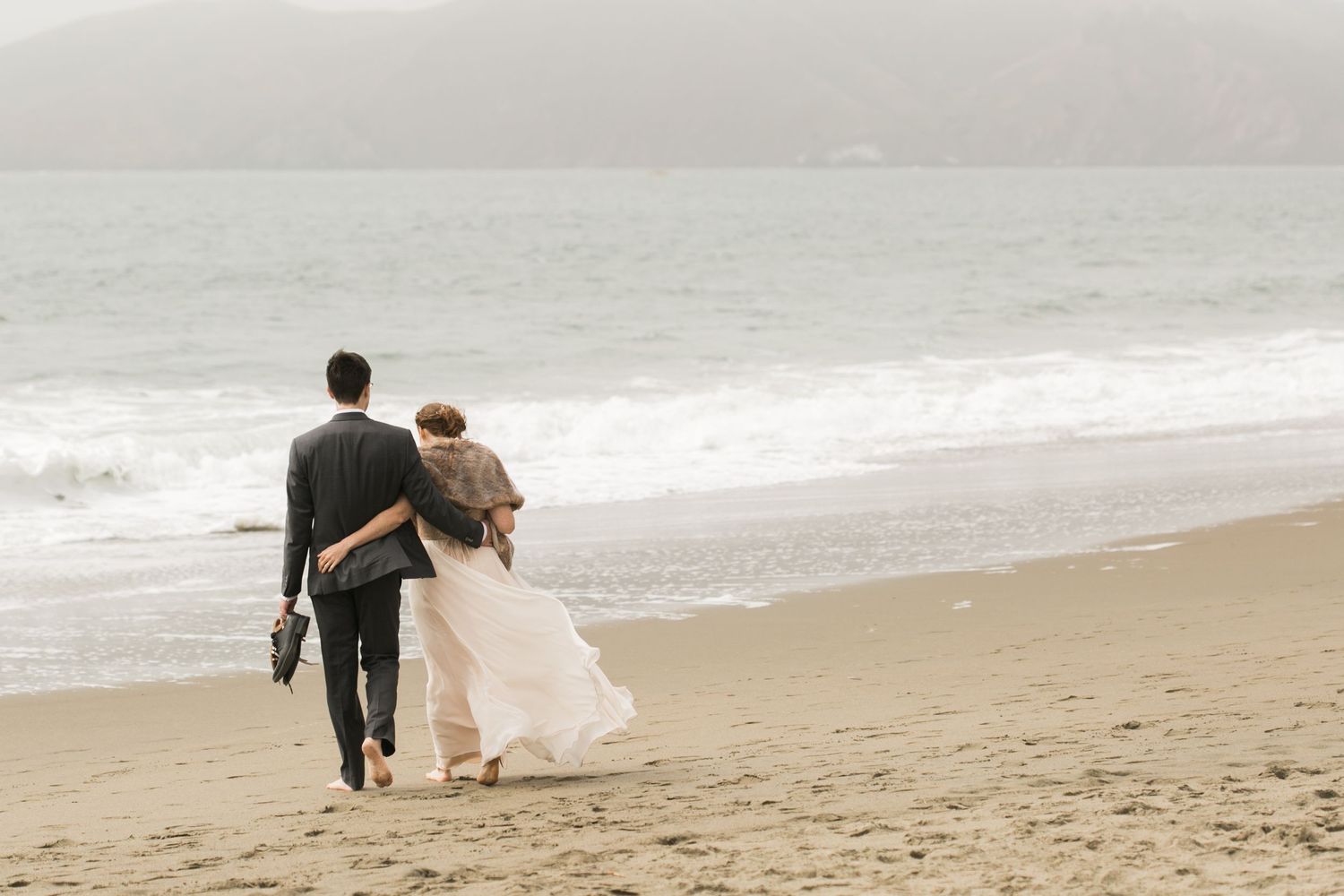 A couple walks hand in hand along a misty beach shoreline in formal attire.