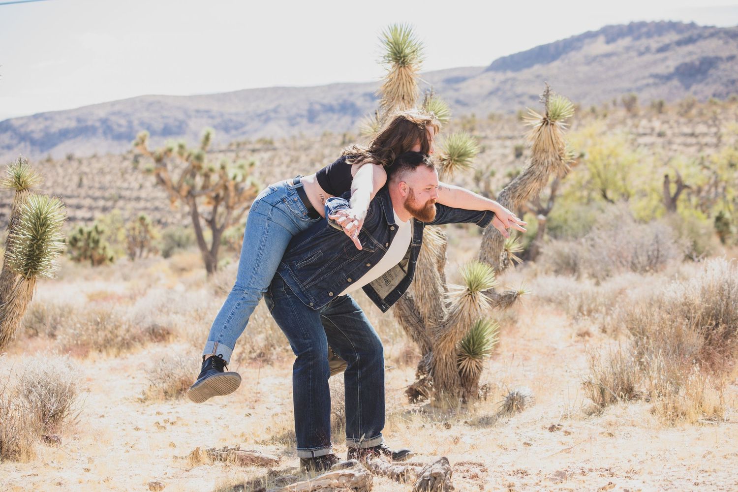 A playful couple in denim enjoying a piggyback ride in a desert landscape.