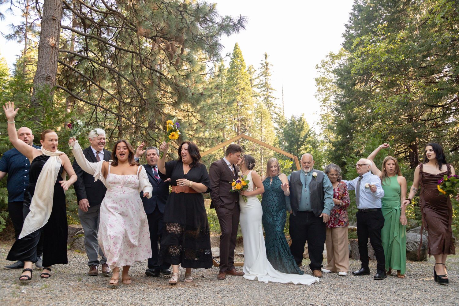 Large wedding group poses for photos in front of tall pine trees during outdoor ceremony.