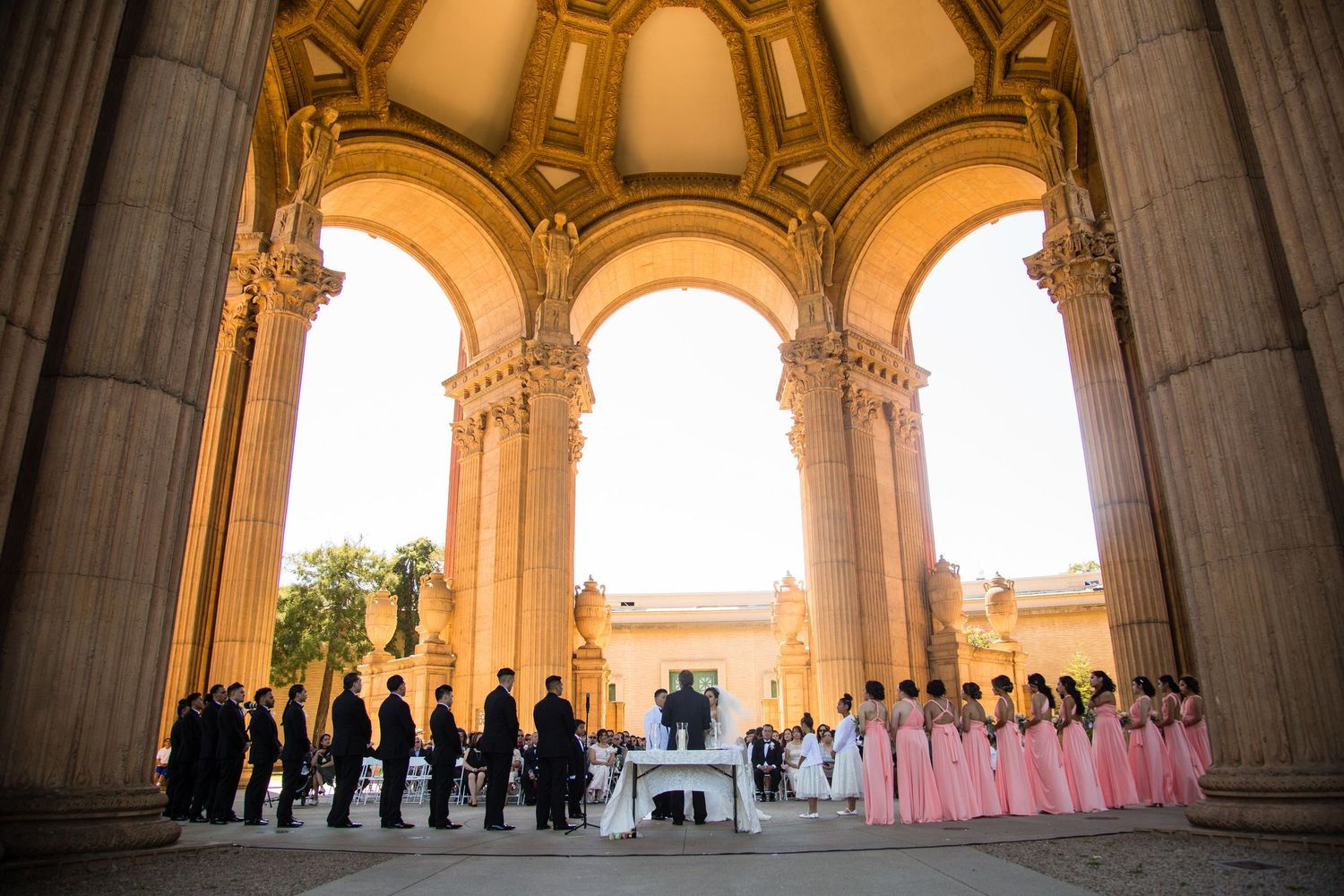 Wedding ceremony under grand arched architecture with wedding party in pink and black attire.