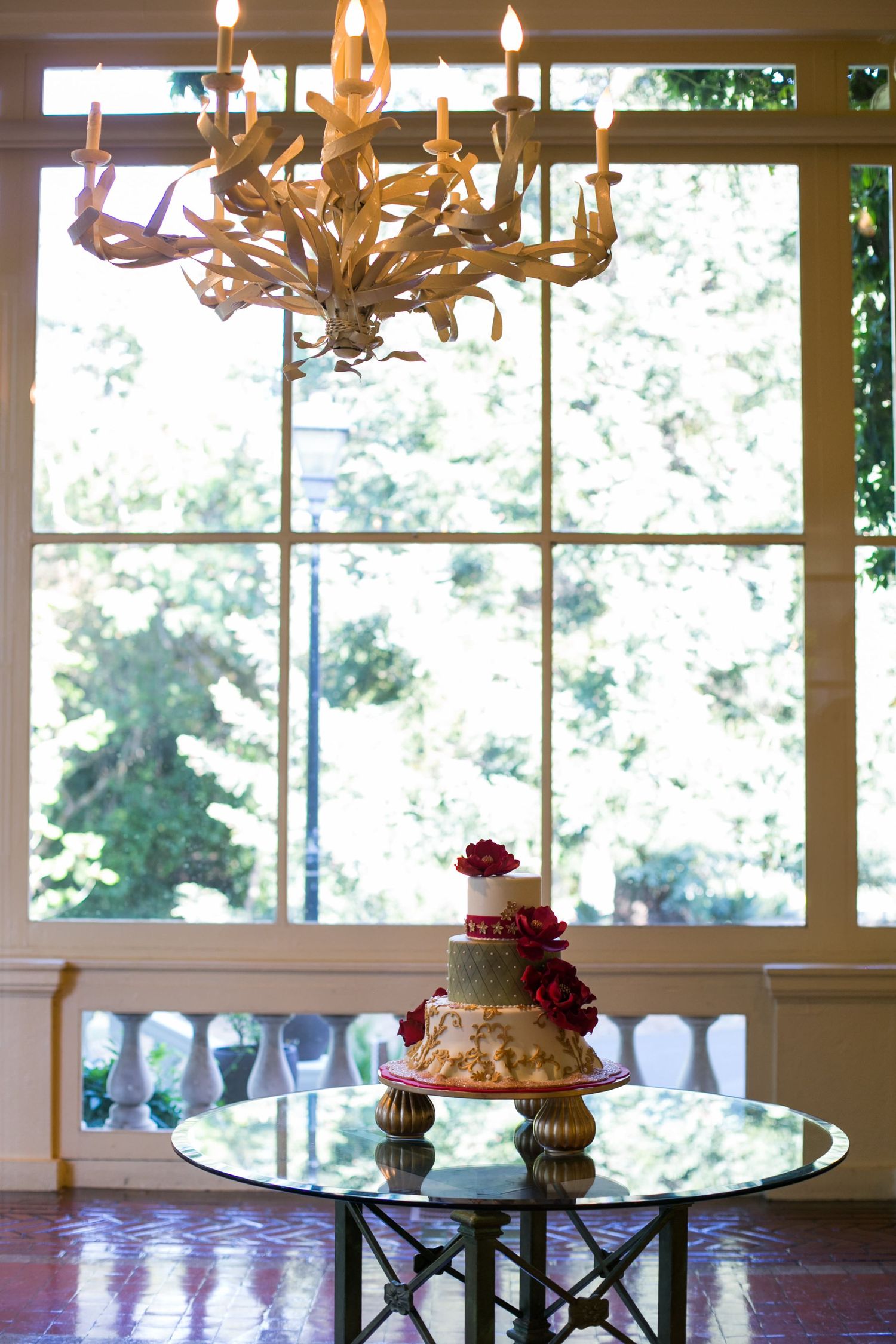 Elegant wedding cake displayed under ornate chandelier near large windows with natural lighting.
