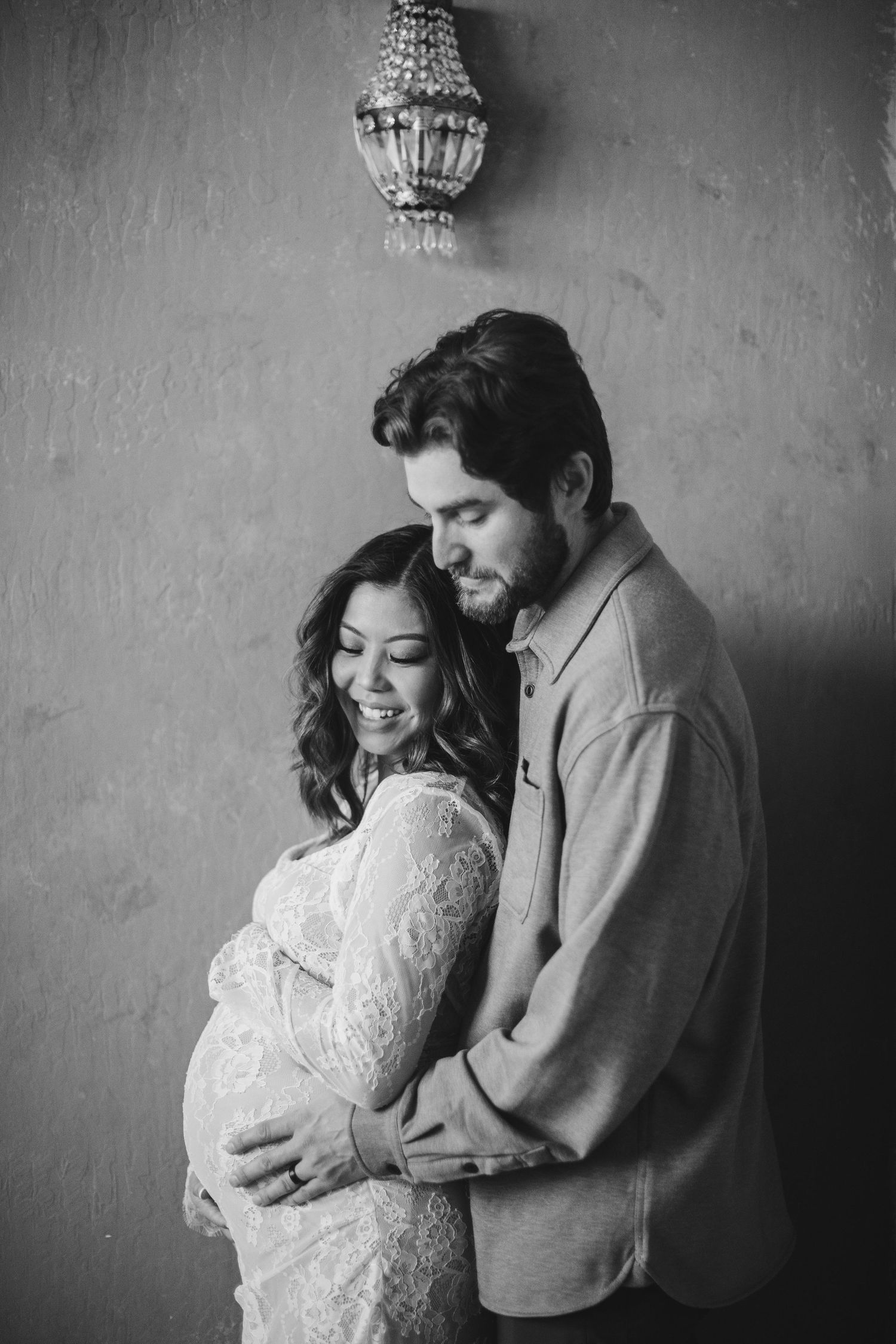 Black and white maternity photo of couple embracing under vintage chandelier.