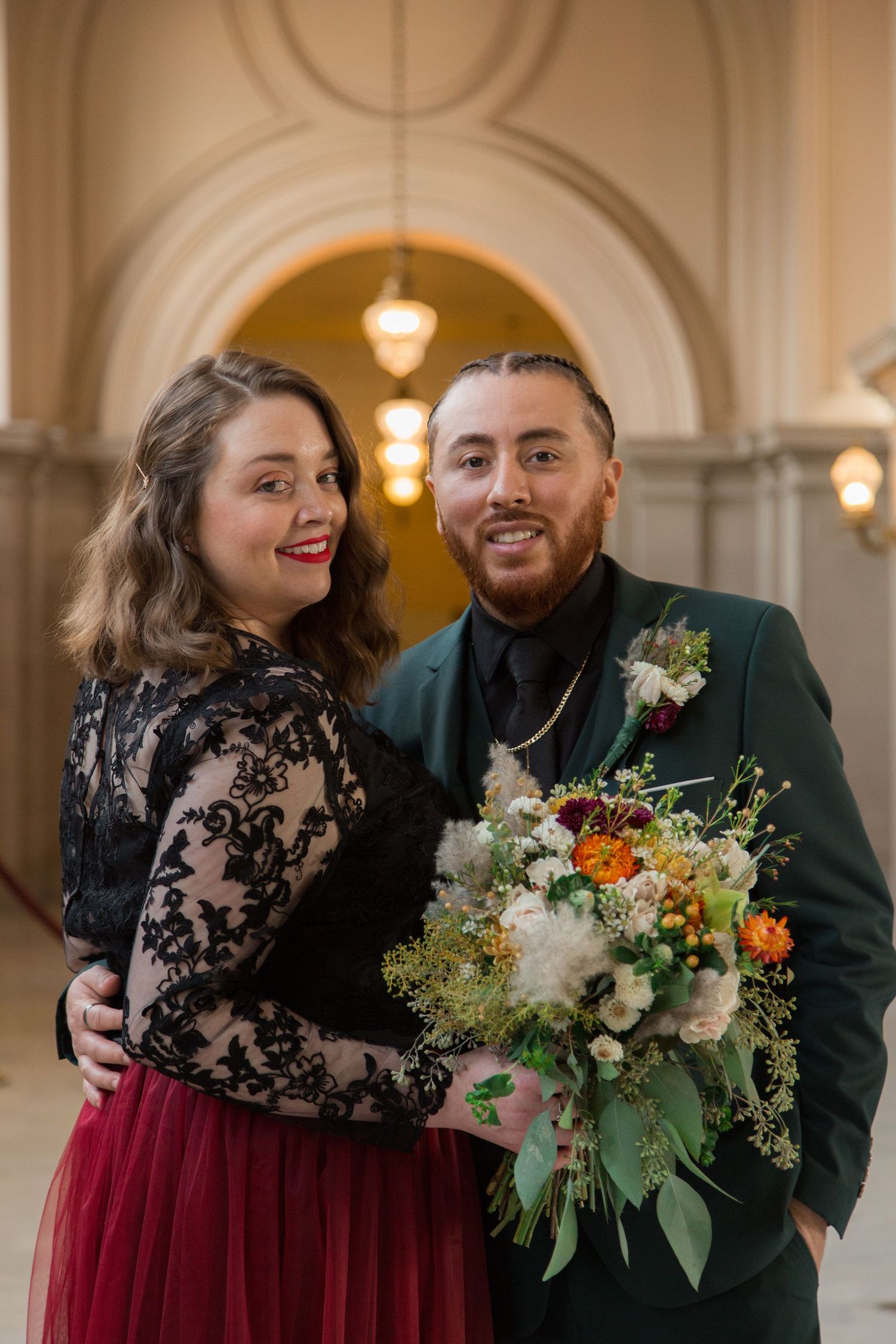 Wedding couple shares candid moments in a grand hall with bride in burgundy skirt and lace top holding colorful bouquet.