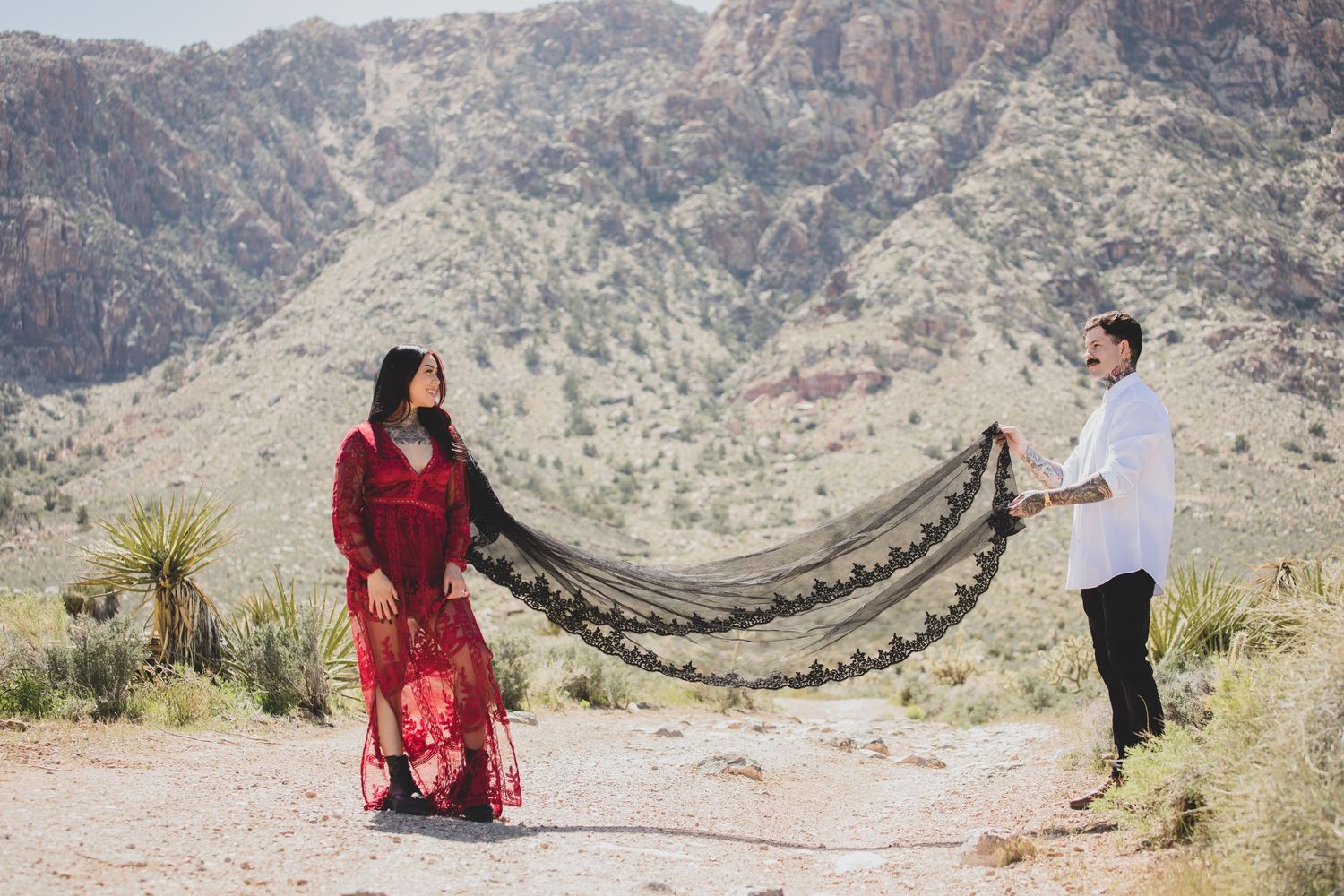 Couple holds black lace train of red dress against desert mountain backdrop.