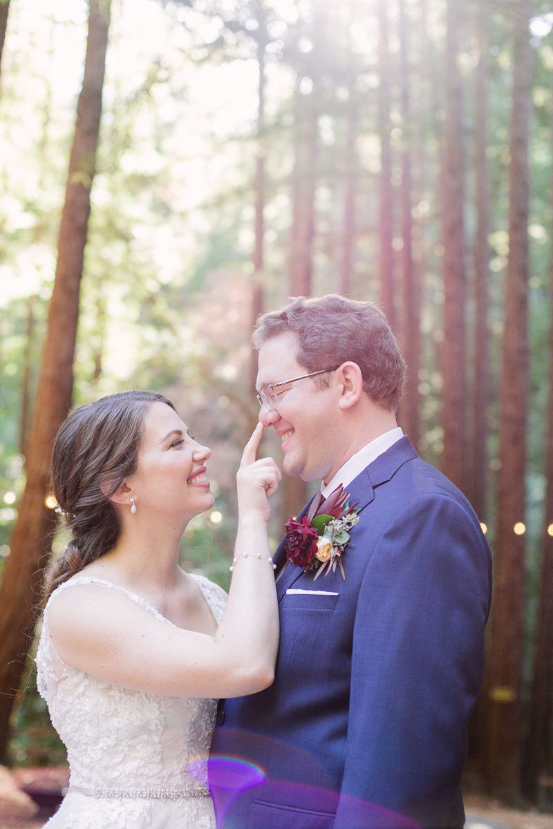 A couple shares joyful laughter together in a sunlit forest setting during a wedding celebration.
