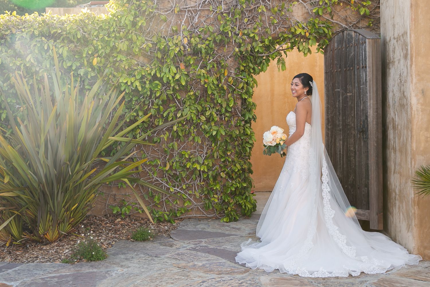 Beautiful wedding gown with lace train against Spanish-style stucco wall and garden setting.
