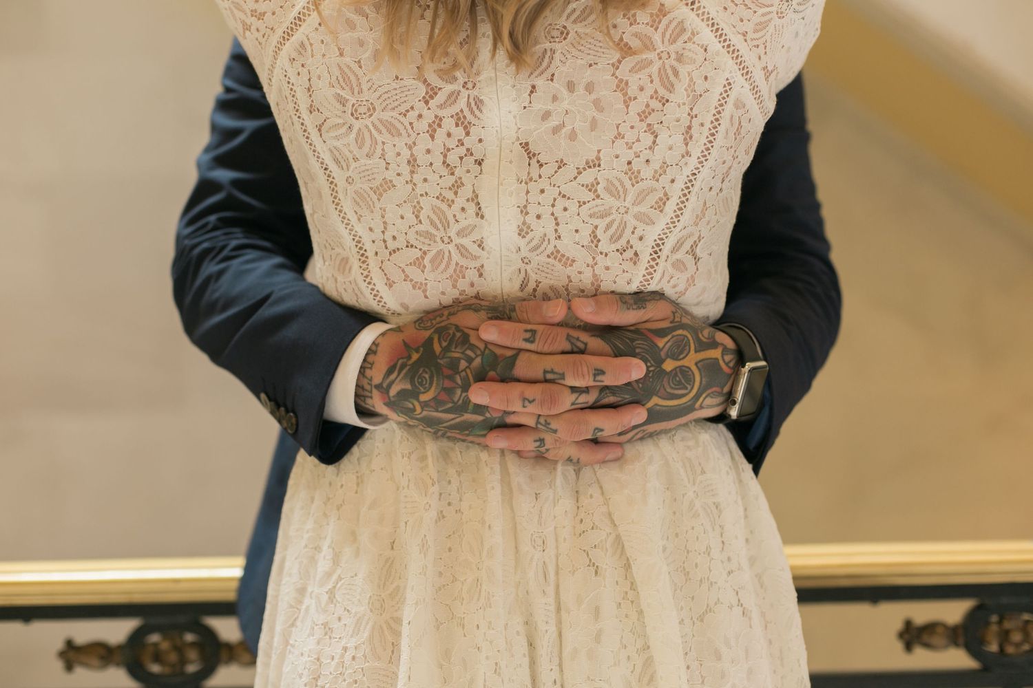 Tattooed hands embrace around waist of white lace wedding dress.