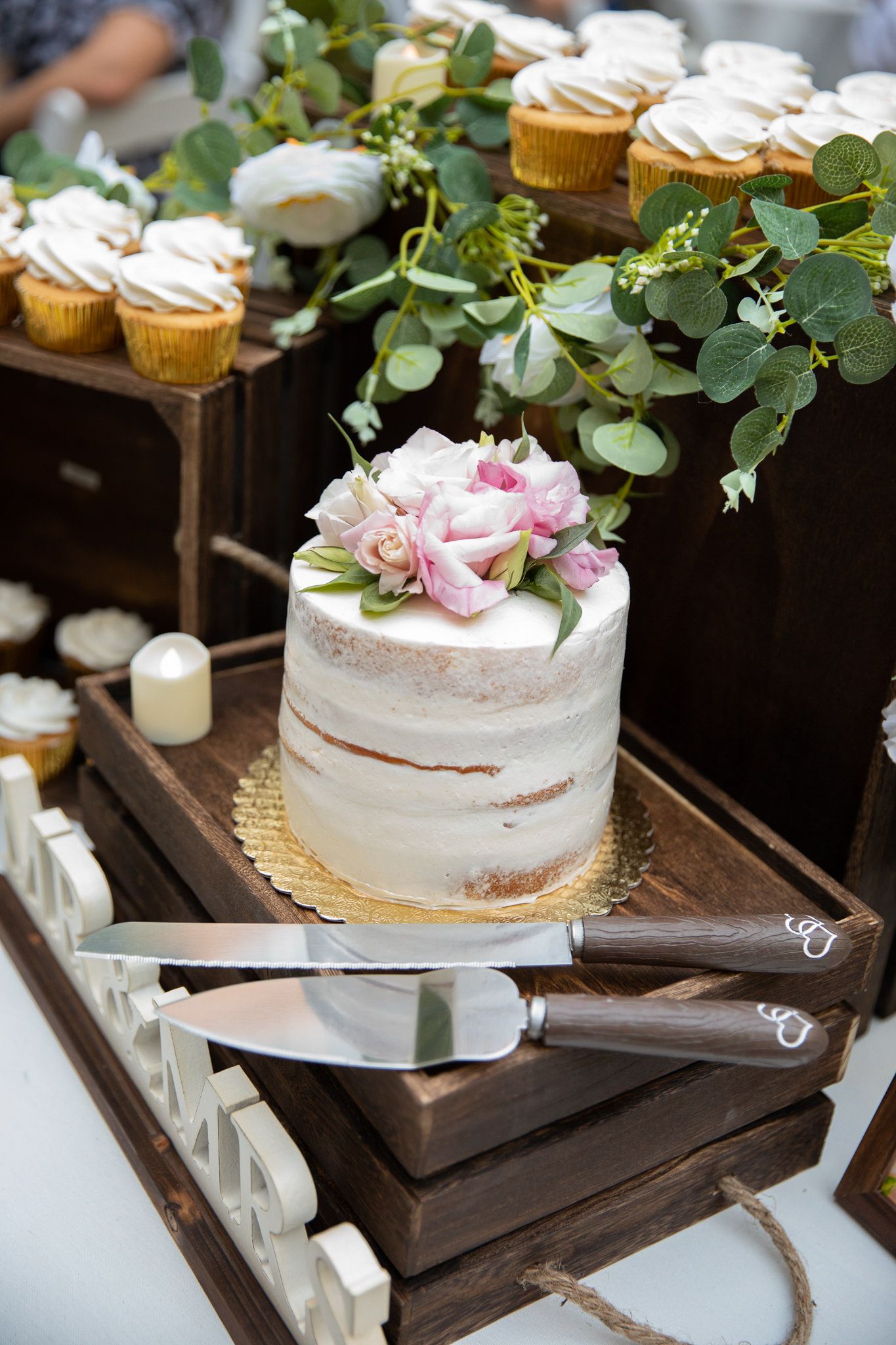 Semi-naked wedding cake decorated with pink roses and eucalyptus alongside cupcakes on wooden display.