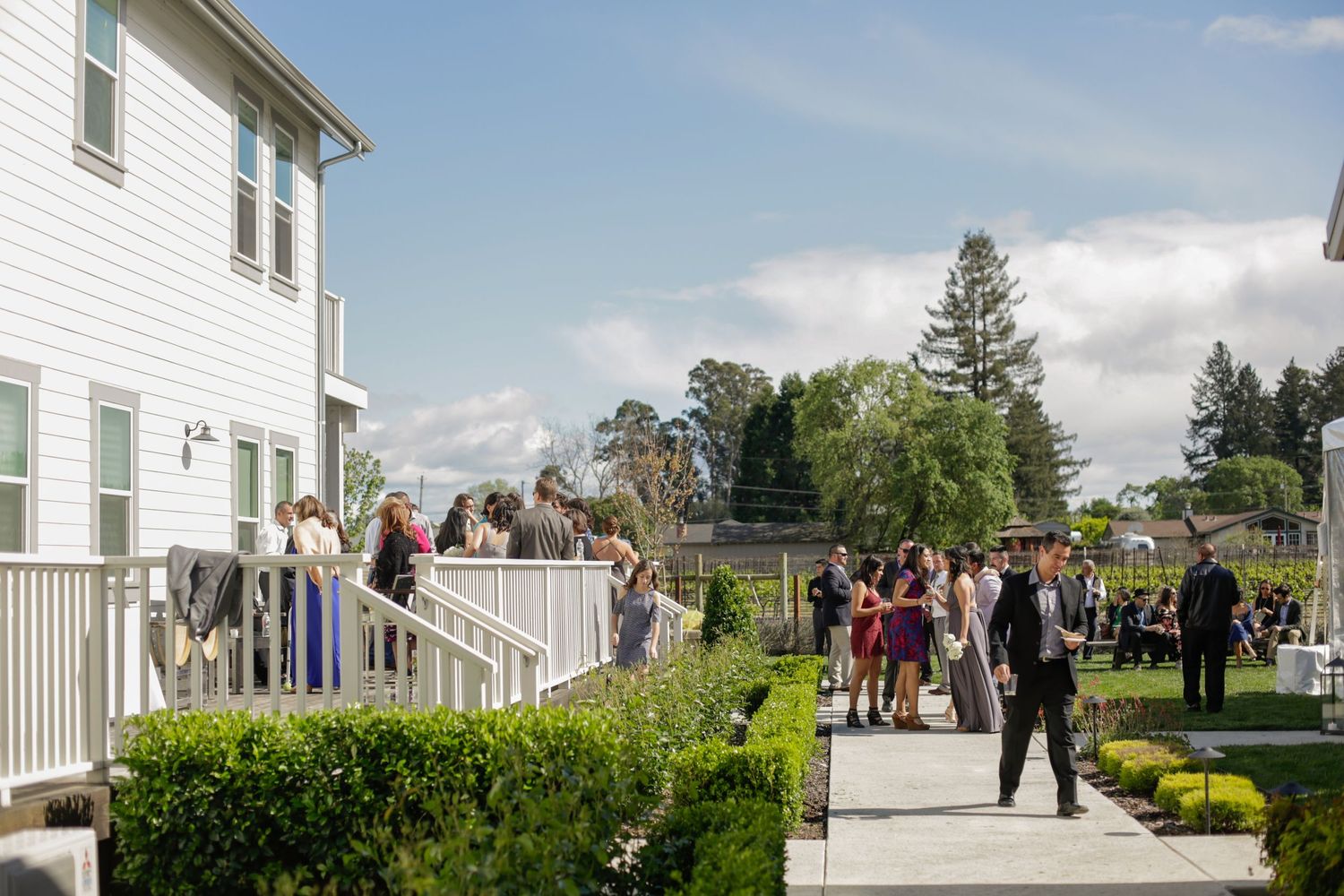 Wedding guests gather on a white porch and walkway surrounded by landscaping on a sunny afternoon.