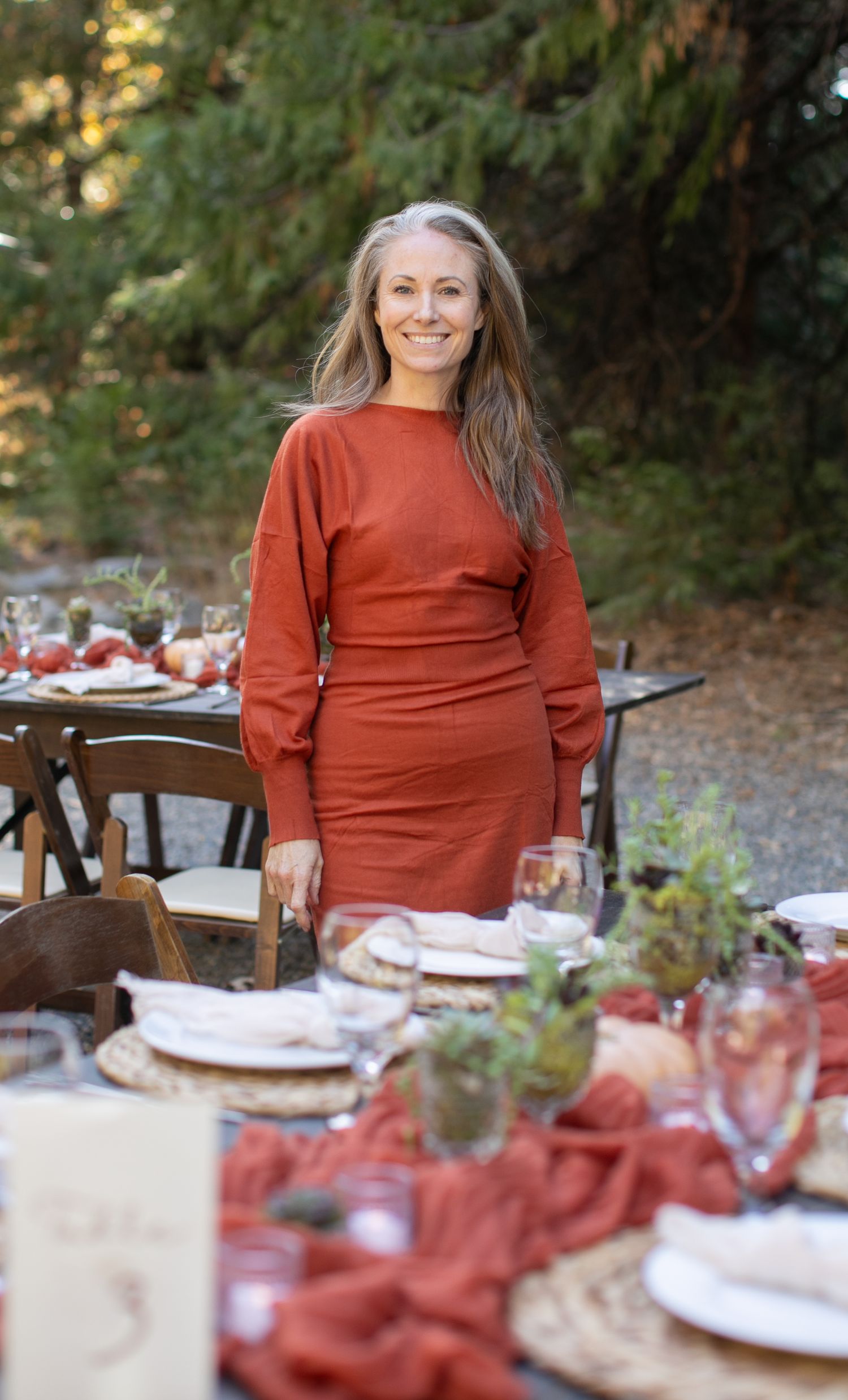 Person in rust-colored sweater dress standing by elegant outdoor table setting in forest.