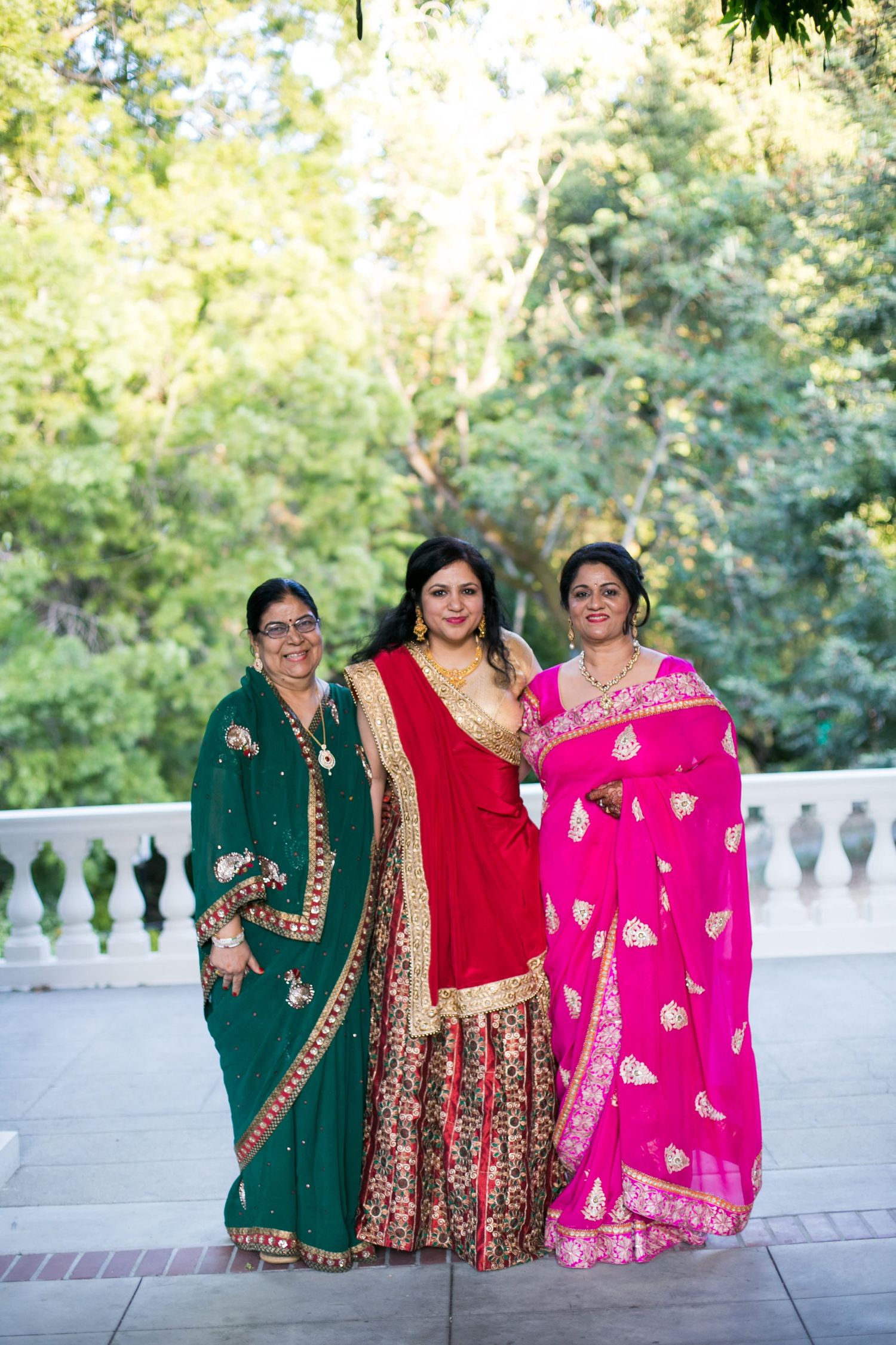 Three women in vibrant traditional Indian saris of green, red, and pink pose outdoors.