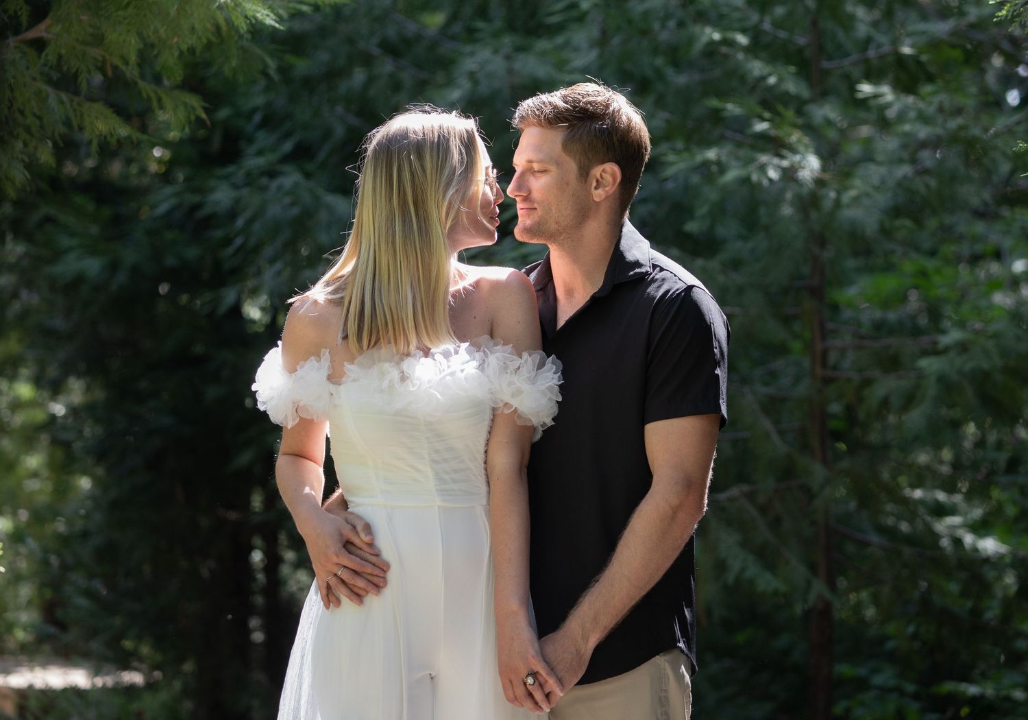 A romantic outdoor engagement photo with a couple in white dress and black shirt surrounded by greenery.