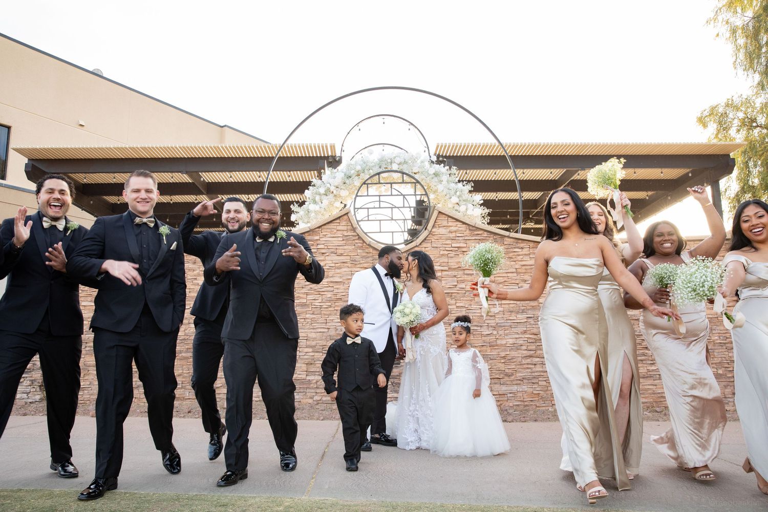 Wedding party jumps joyfully for a fun photo under an outdoor arch.