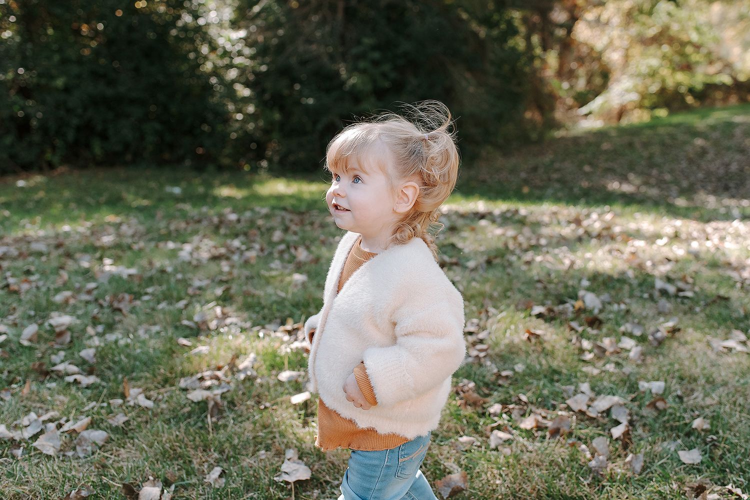 A toddler in jeans and a cream sweater stands in a sunlit garden with dappled light through the trees.