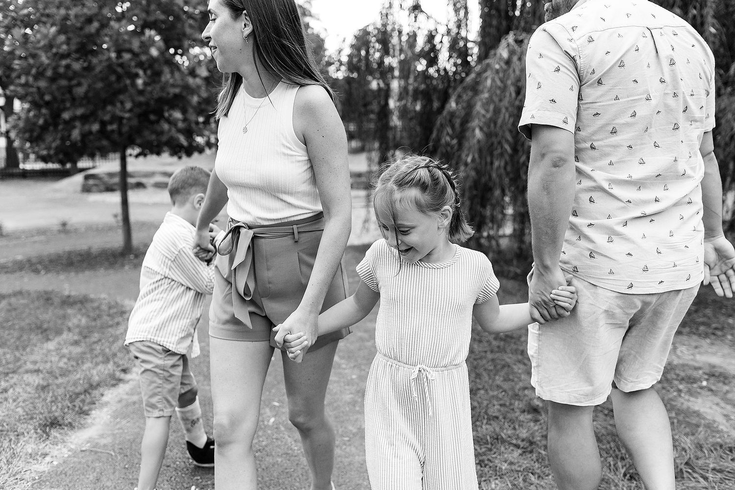 Black and white outdoor photo showing a family holding hands while walking together in a park setting during summer.