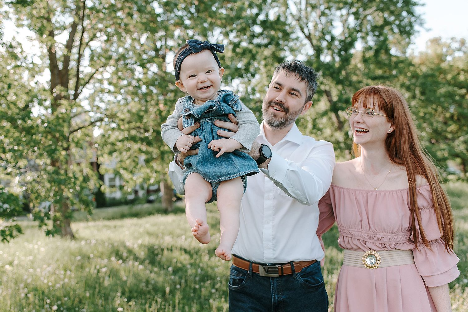 A family stands together outdoors in a sunny field with green trees in the background for a portrait session.
