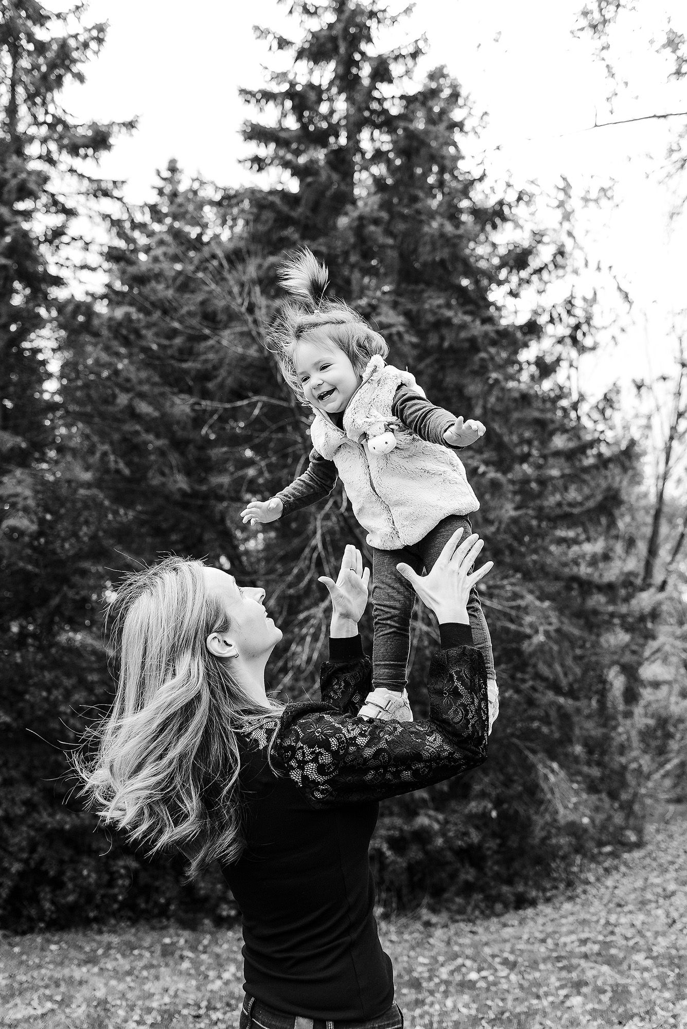 A black and white outdoor photo of a parent lifting a joyful child high in the air with evergreen trees in the background.