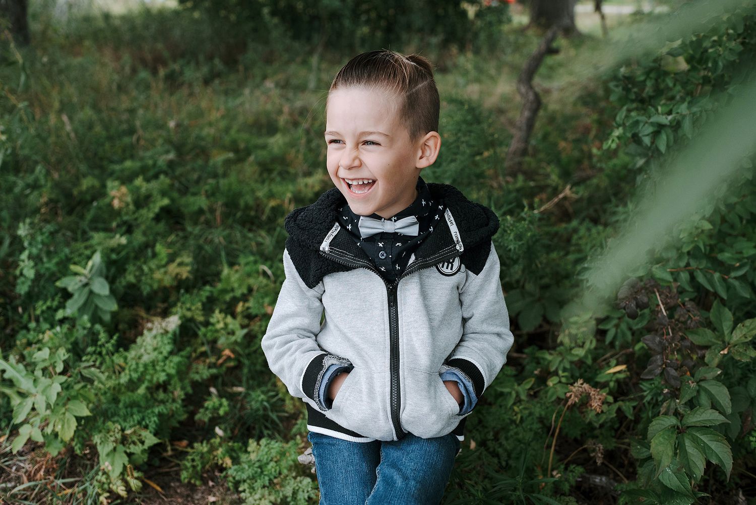 A child wearing a gray and black jacket stands smiling among green foliage in an outdoor setting.