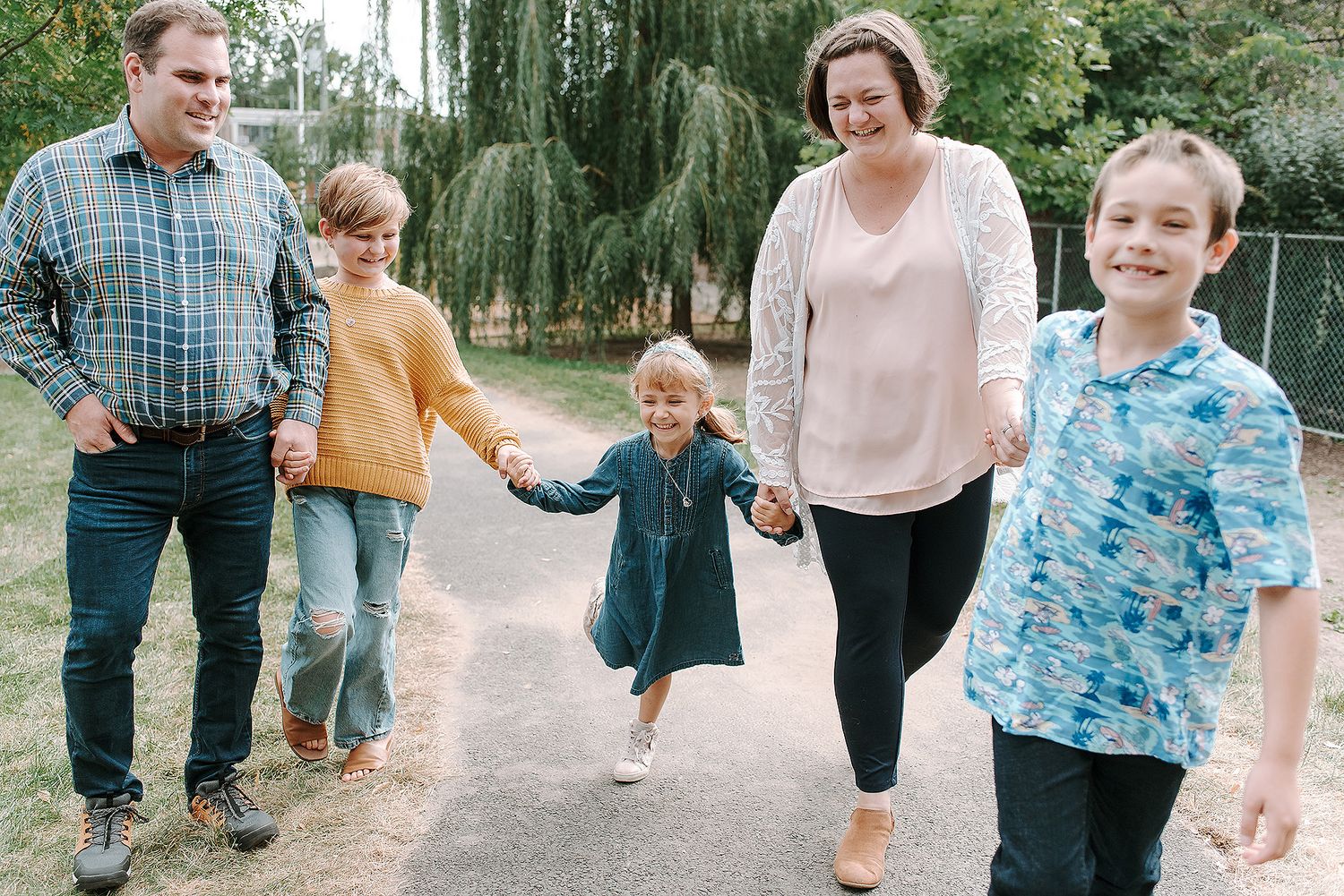 Family members walking together holding hands on a path under willow trees on a sunny day.