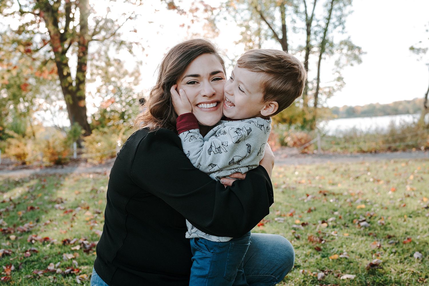 Two people embracing outdoors in autumn with fallen leaves and a lake visible in the background.