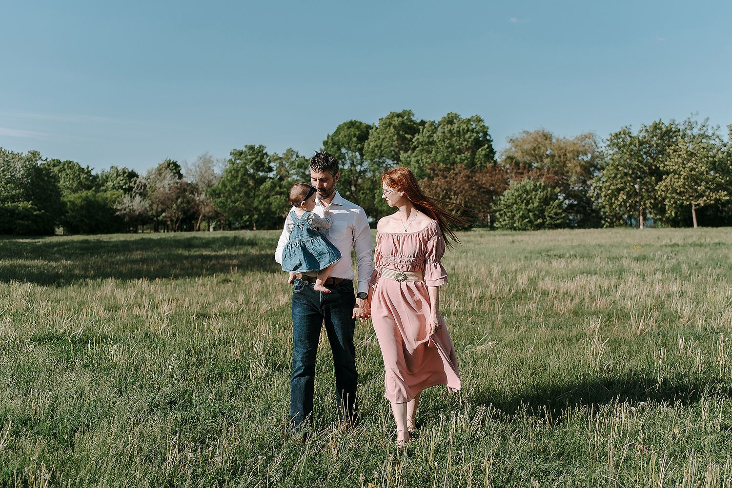 A couple in casual attire stands together in a sunny meadow with trees in the background on a clear day.