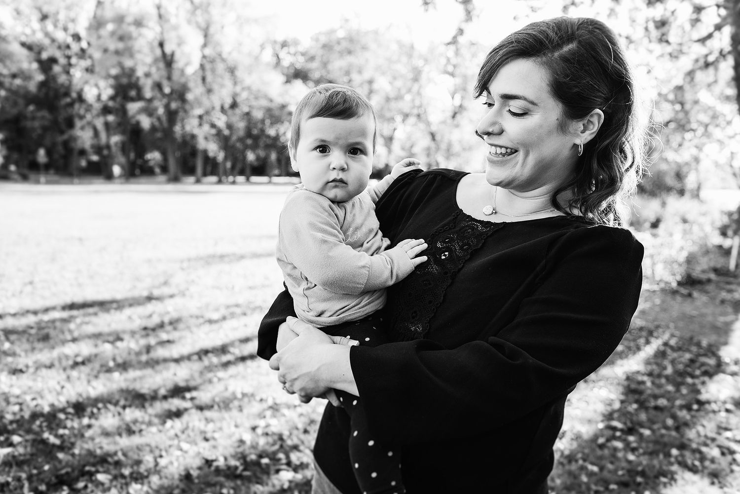 Black and white photo of a mother holding a baby while smiling outdoors in a natural setting.