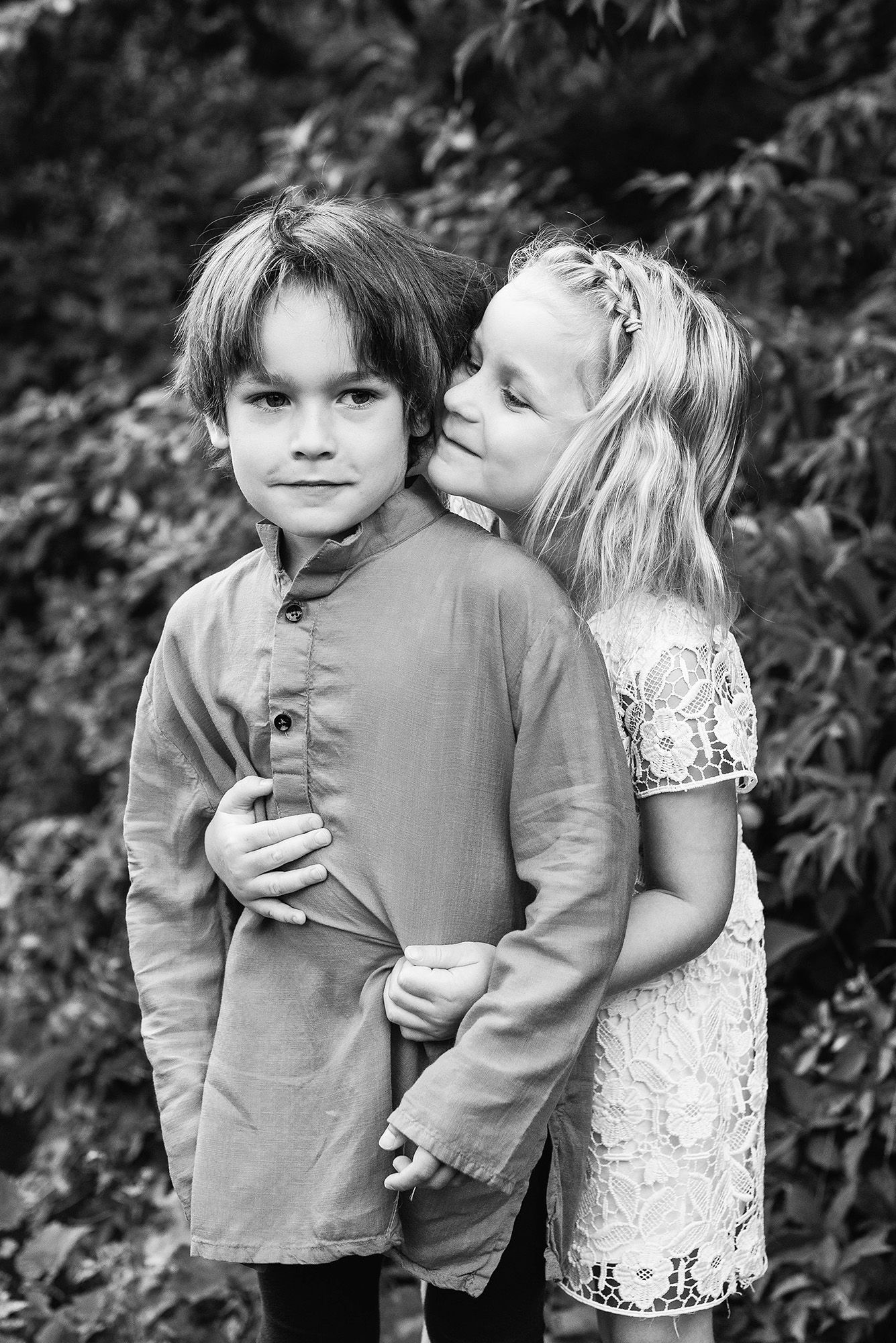 A black and white portrait of two children in casual attire standing close together outdoors near foliage.