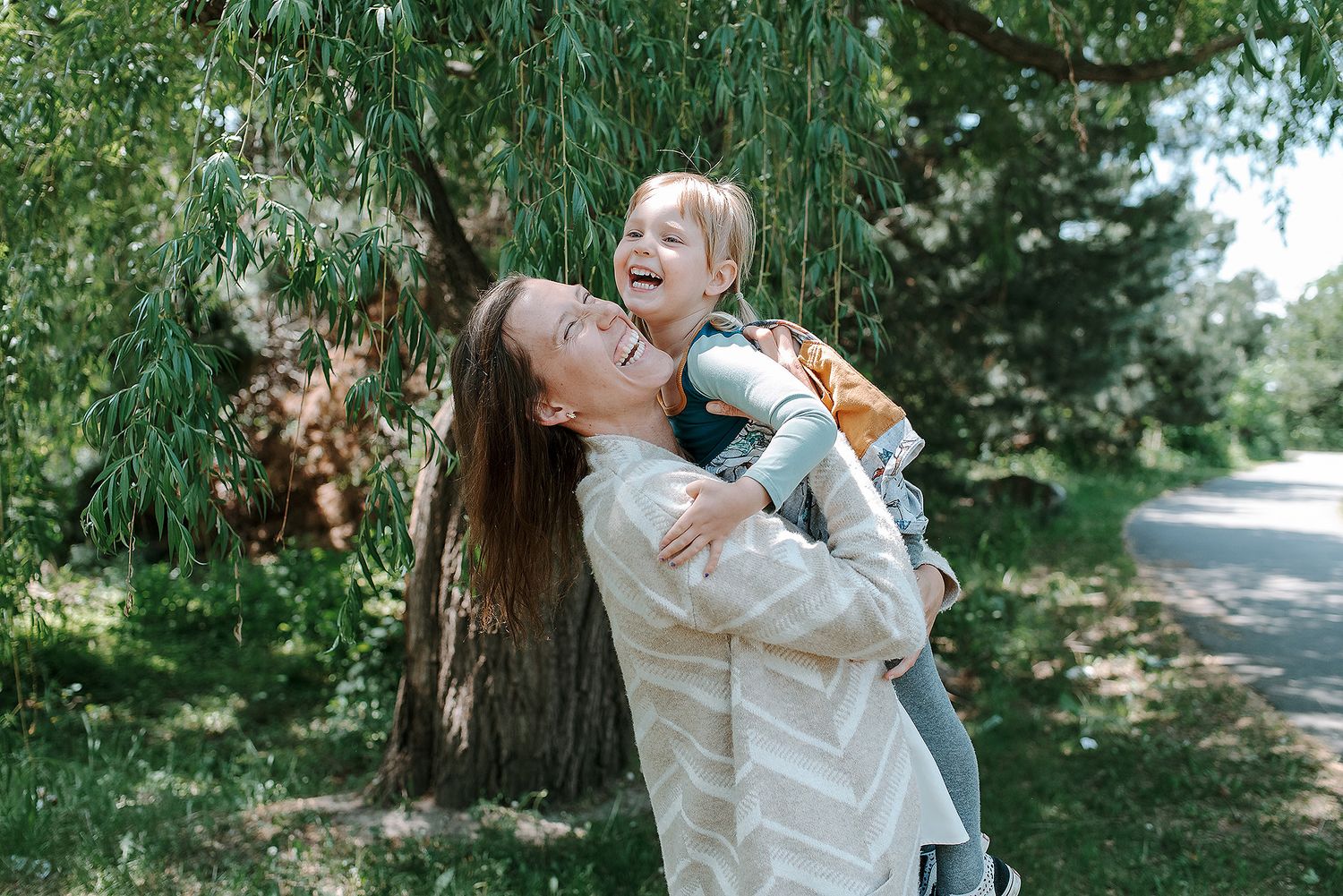 Parent and child share joyful moment playing outdoors on sunny day under willow tree along path.