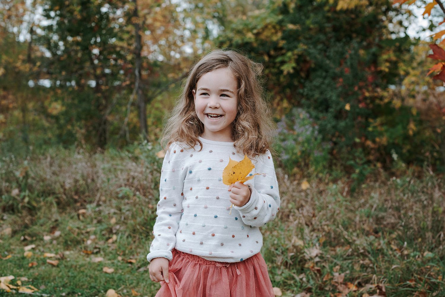 A child wearing a polka dot sweater holds a golden autumn leaf while standing outdoors in nature.