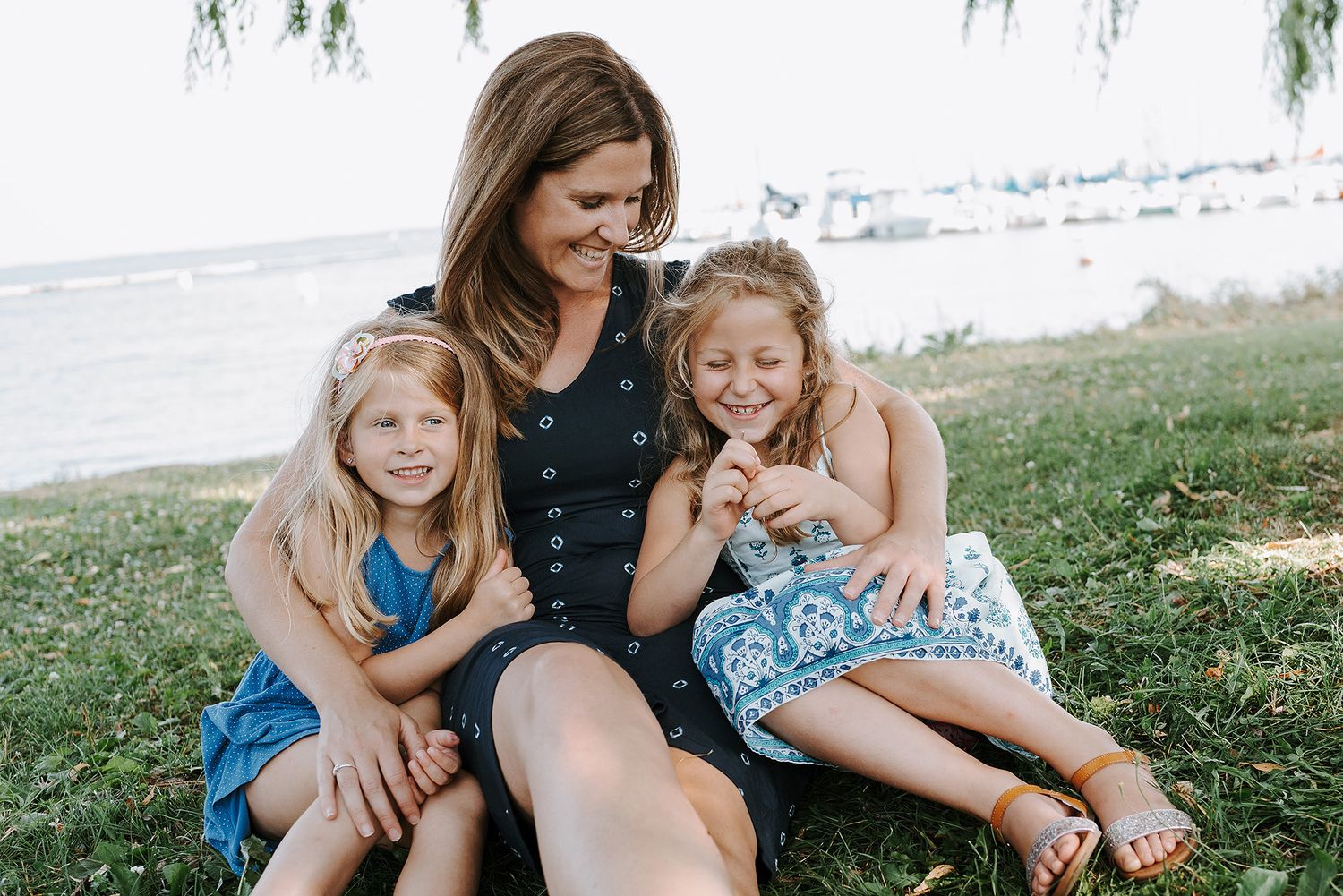 A tender moment shared between family members sitting together on grass near a waterfront during a sunny day.