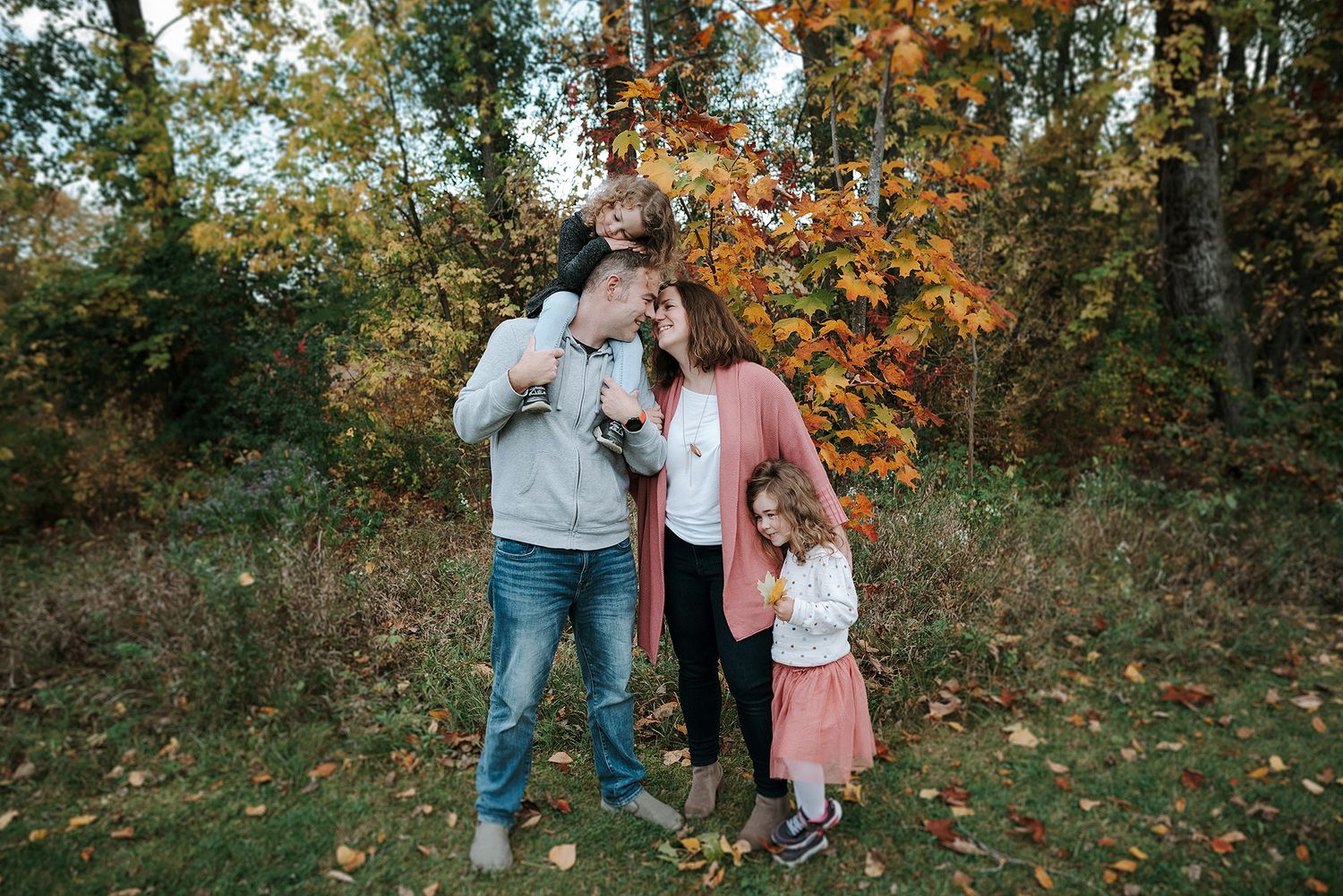 A family with their small dog stands together in autumn woods with colorful fall foliage in the background.