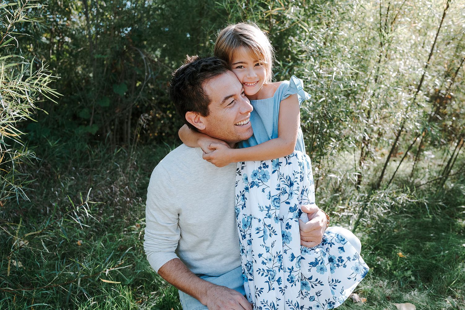 Joyful outdoor portrait of parent and child hugging amidst lush greenery on a sunny afternoon.