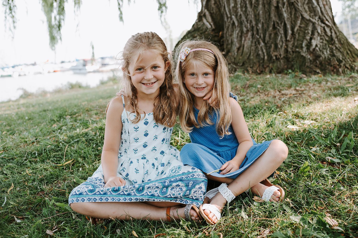 Two children sit close together under a large tree on grass near the waterfront.