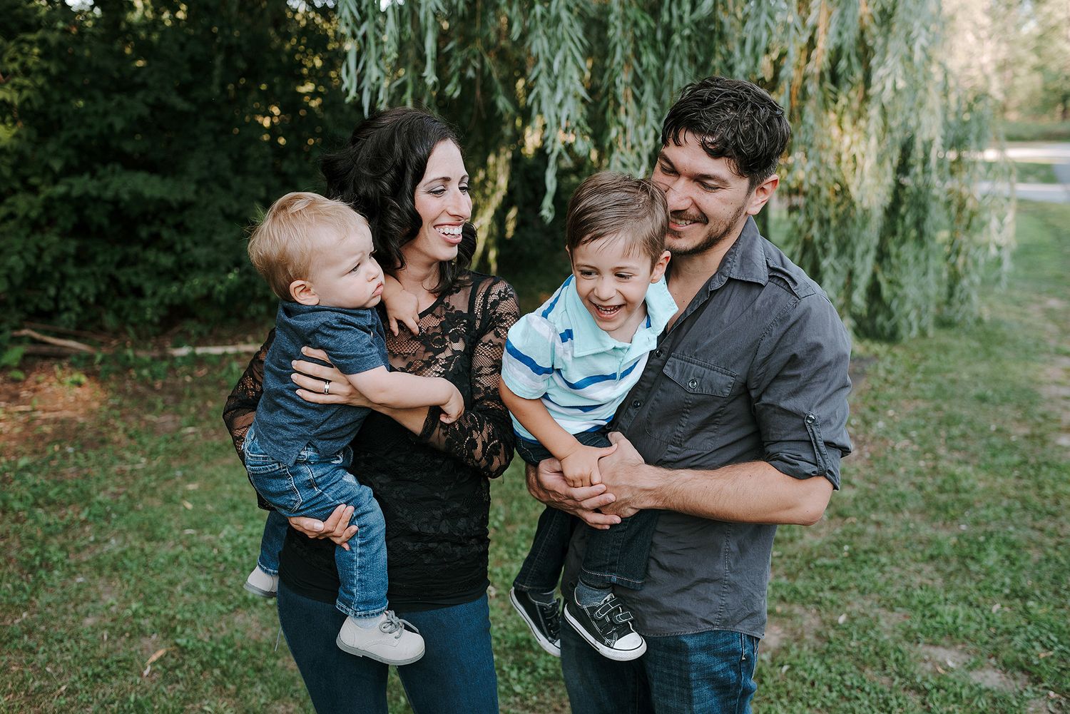 A family laughs and plays together beneath a weeping willow tree in an outdoor setting.