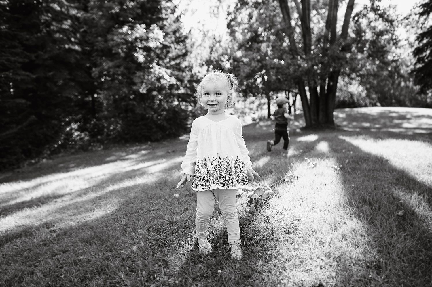 Black and white photo of a young child wearing a floral dress standing on a sunlit outdoor path among trees.