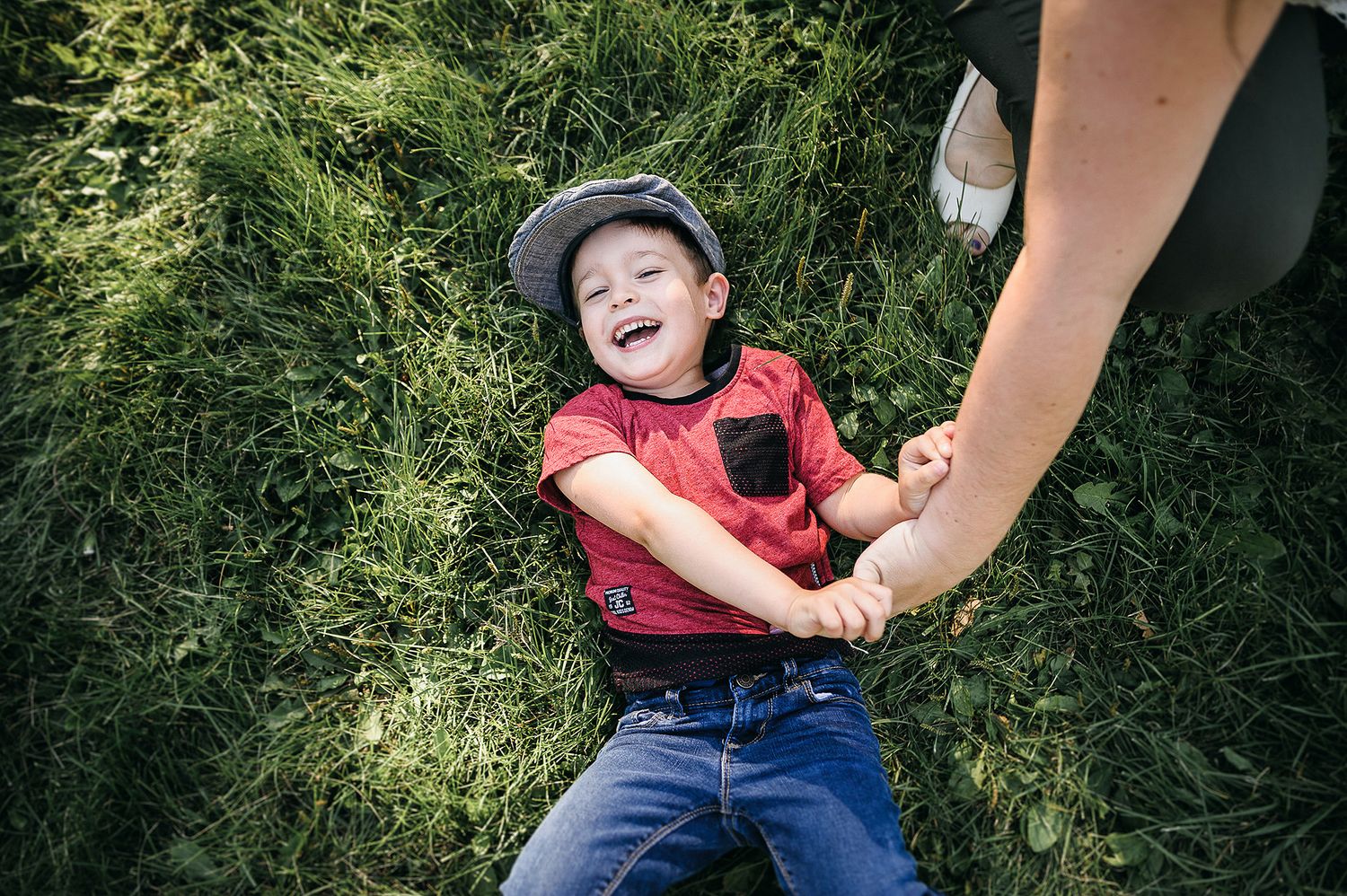 Joyful child wearing a red vest and cap lies in green grass while holding an adult's hand.