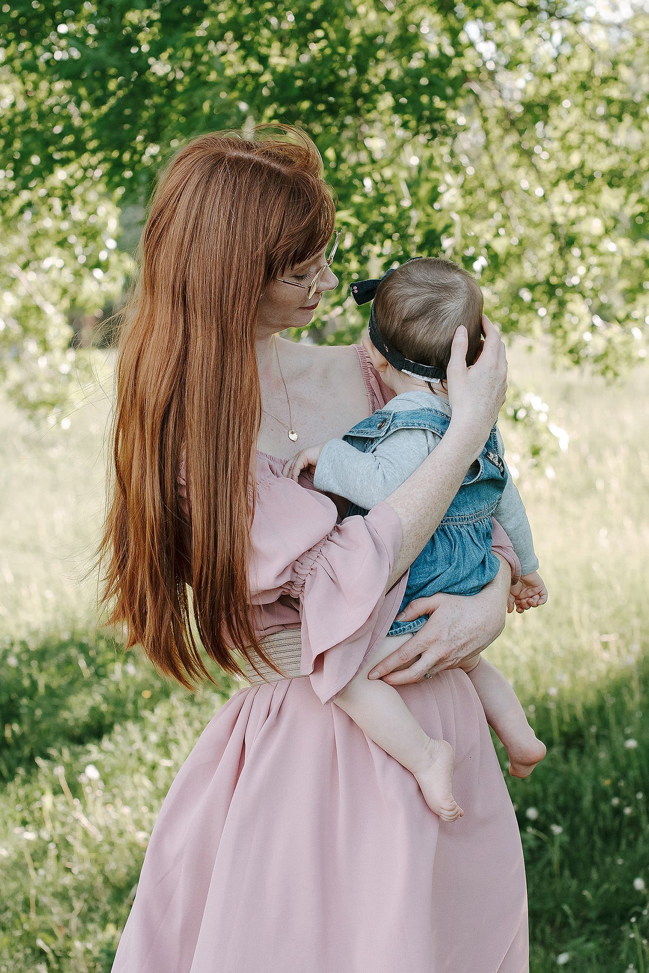 A woman in a pink dress holds a baby wearing blue clothing in a sunny outdoor setting among green foliage.