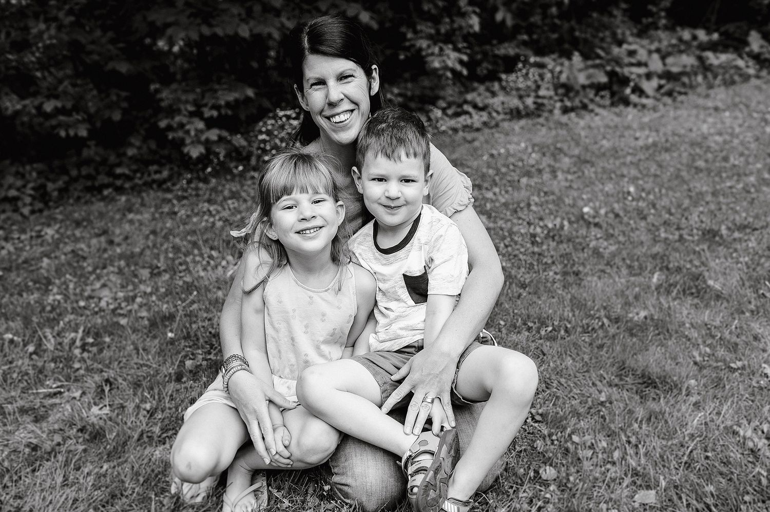 A black and white photograph shows three people sitting together outdoors on grassy ground during summertime.