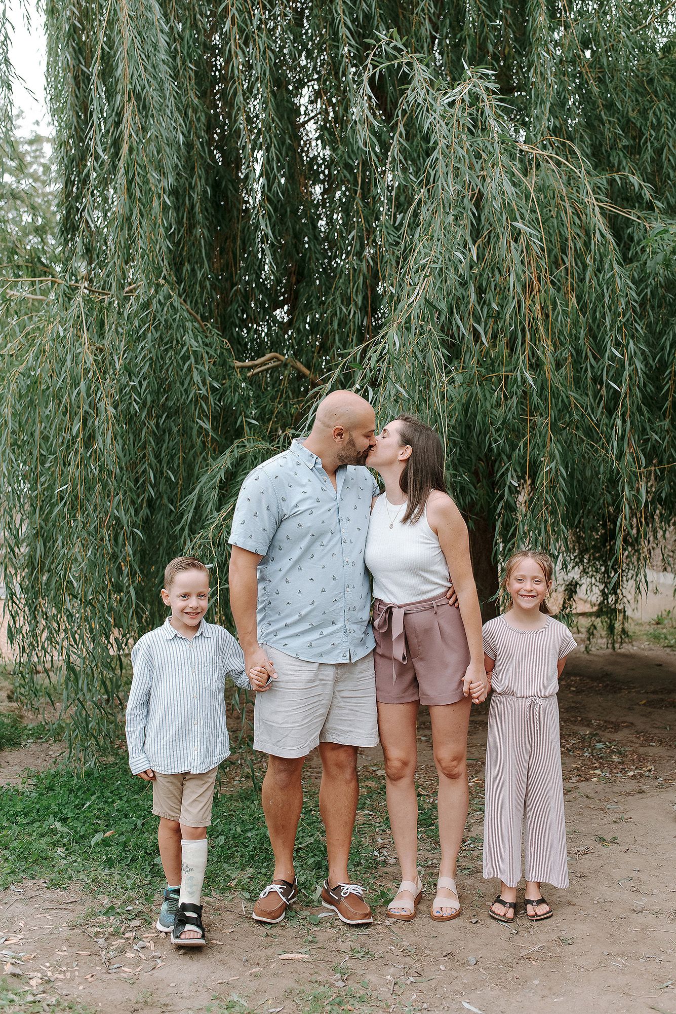 Family stands together on dirt path under drooping branches of a majestic willow tree on sunny day.