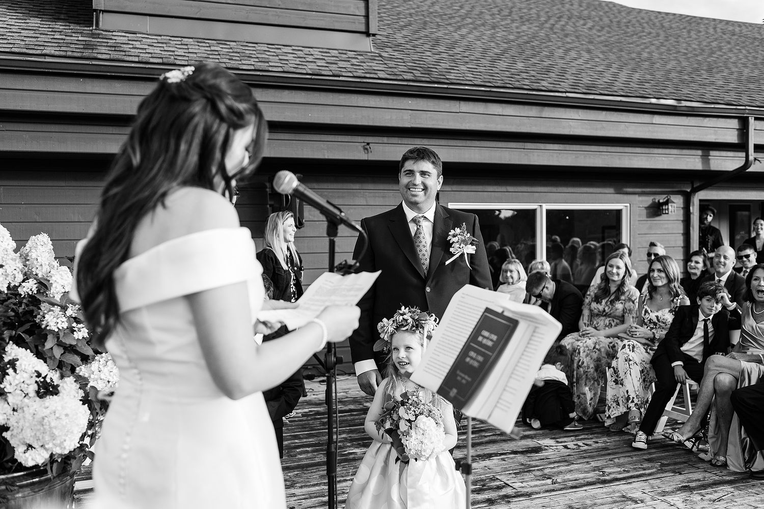 Black and white photo of a wedding ceremony on an outdoor deck with guests seated and beautiful floral arrangements. Grand Lodge.